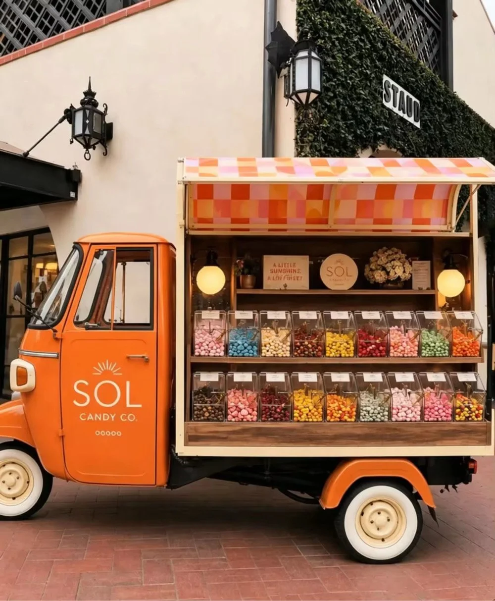 Orange candy truck with the logo 'SOL CANDY CO.' parked outside a building. The truck has a colorful checkered awning and is filled with various colorful candies in transparent containers.