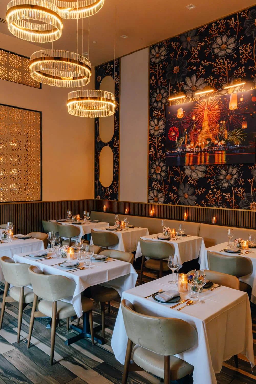 Elegant restaurant dining area with white tablecloths, ambient candle lighting, and neatly arranged glassware and silverware. Decorative wall art featuring Paris with fireworks, floral wallpaper, modern hanging light fixtures, and comfortable upholstered chairs.