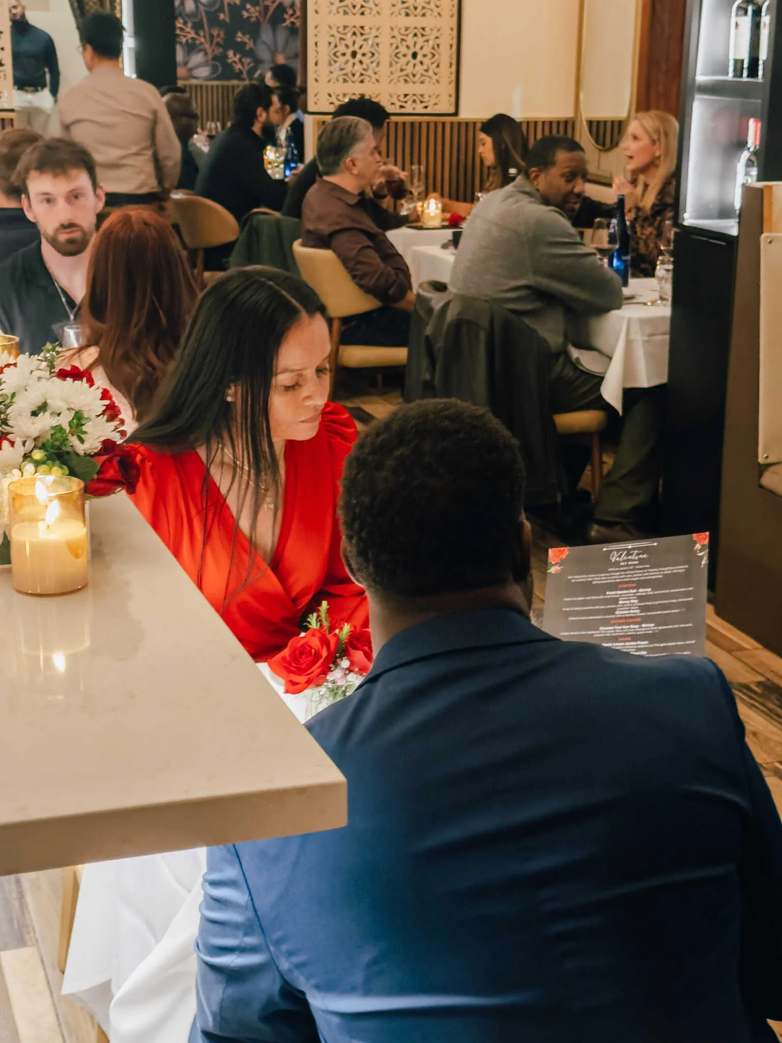 People dining at a wedding reception or celebration, seated at tables with floral centerpieces and candles, engaging in conversations in a modern restaurant or banquet hall.