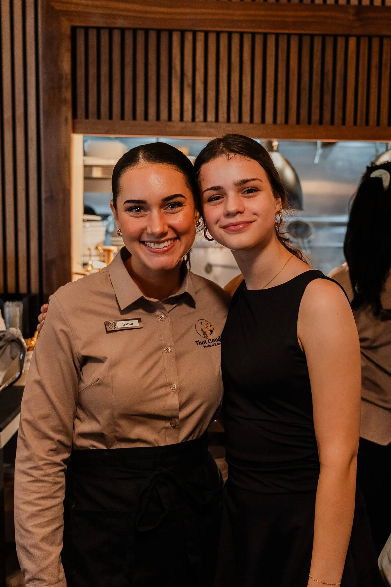 Two women smiling, one wearing a beige uniform with a name tag that says 'Sarah' and a logo for Thai Candy, the other in a sleeveless black top, standing in a restaurant kitchen.