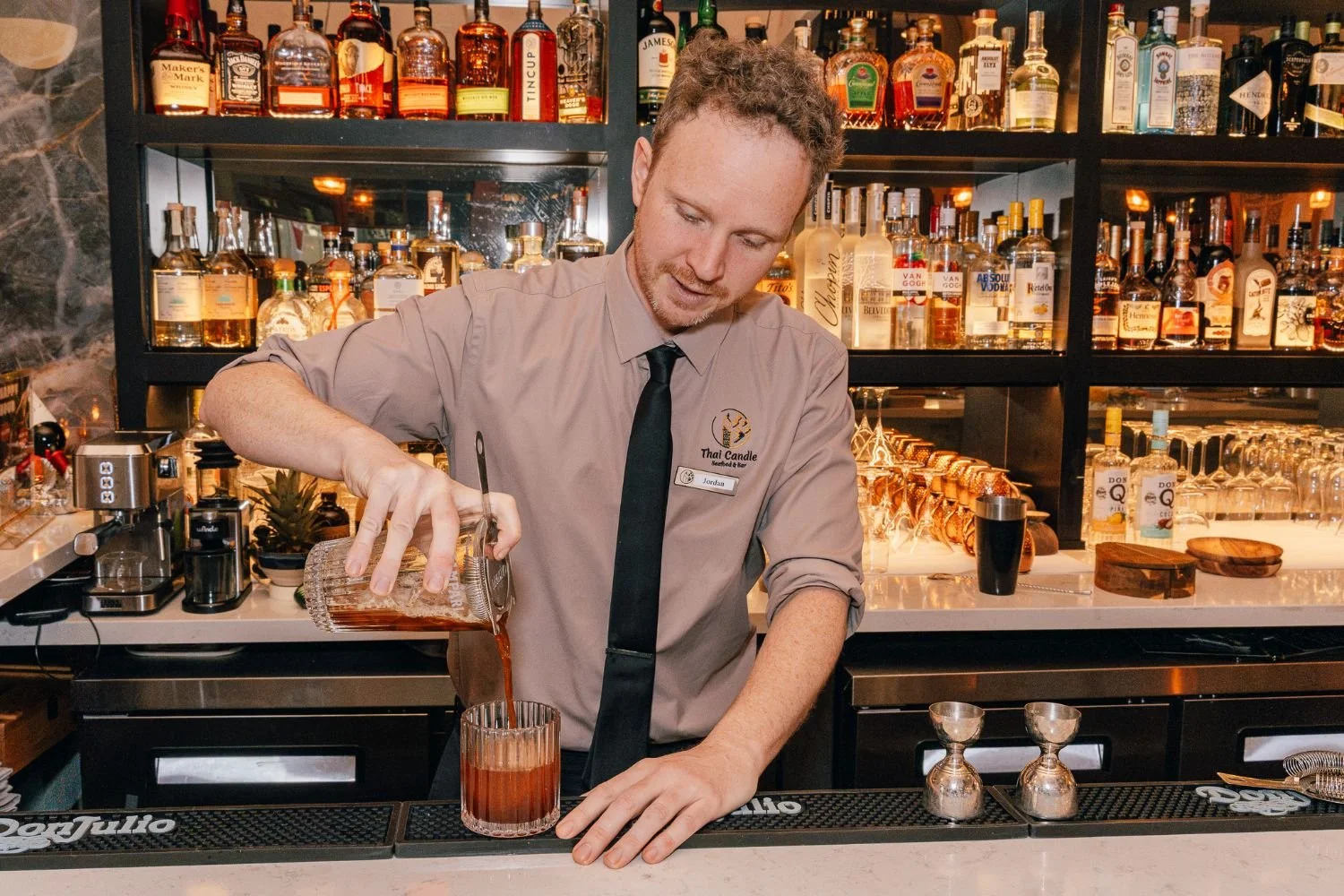 A bartender pouring a drink into a glass at a bar with shelves of liquor bottles behind him.