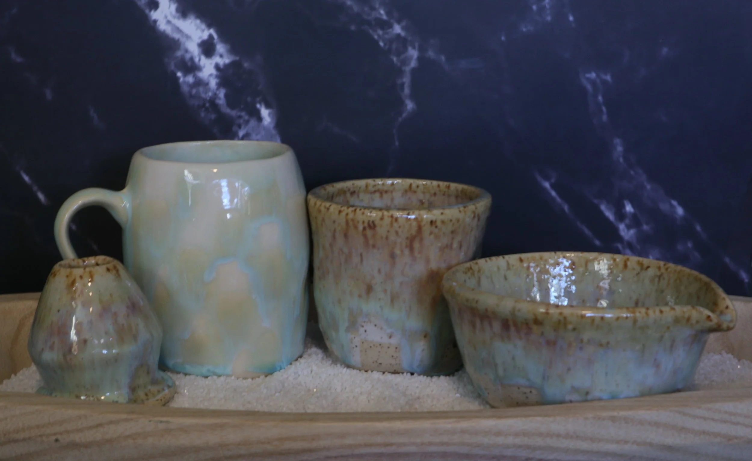 Various ceramic mugs and cups displayed on a wooden shelf with a dark, textured background.