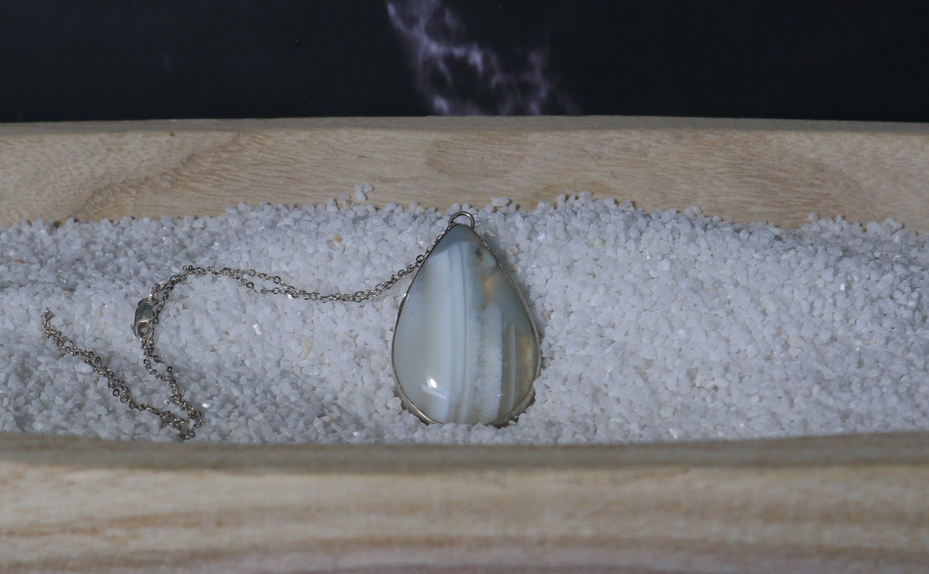 A teardrop-shaped pendant with a striped pattern on a chain, resting on white gravel inside a wooden box.