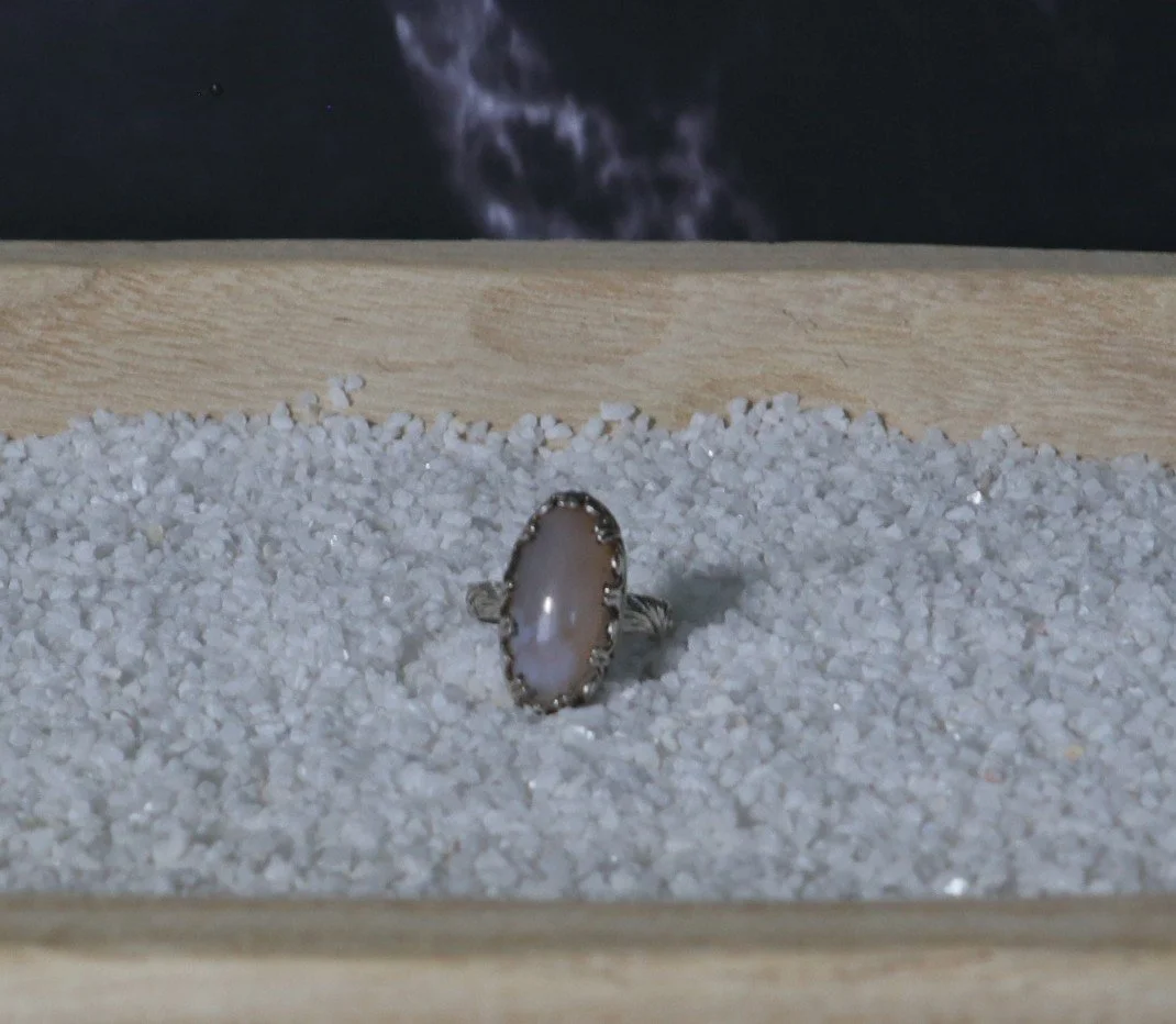 A ring with a large oval stone set in a silver band, displayed on white sands in a wooden box.
