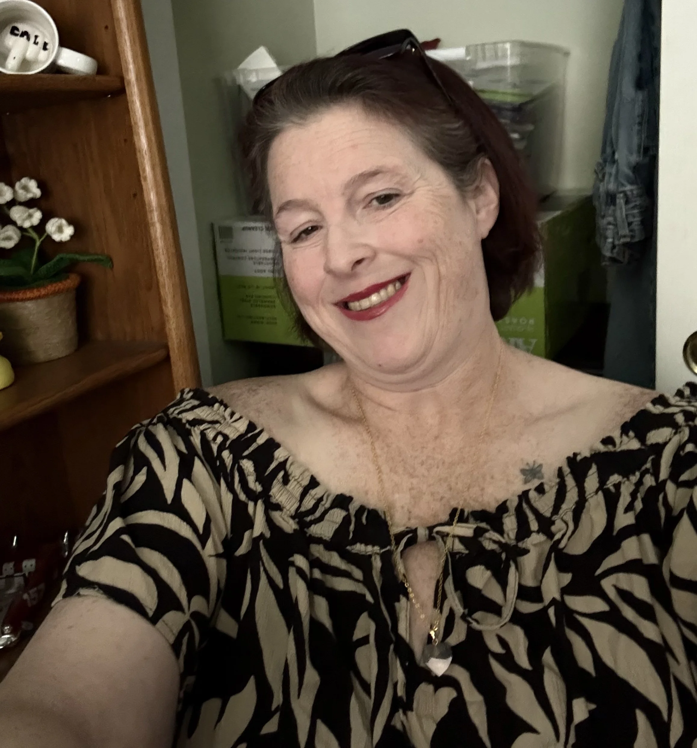A woman with short auburn hair, wearing glasses on her head, a patterned black and beige animal print top, a gold necklace, and red lipstick, smiling in a room with wooden shelves and storage containers in the background.
