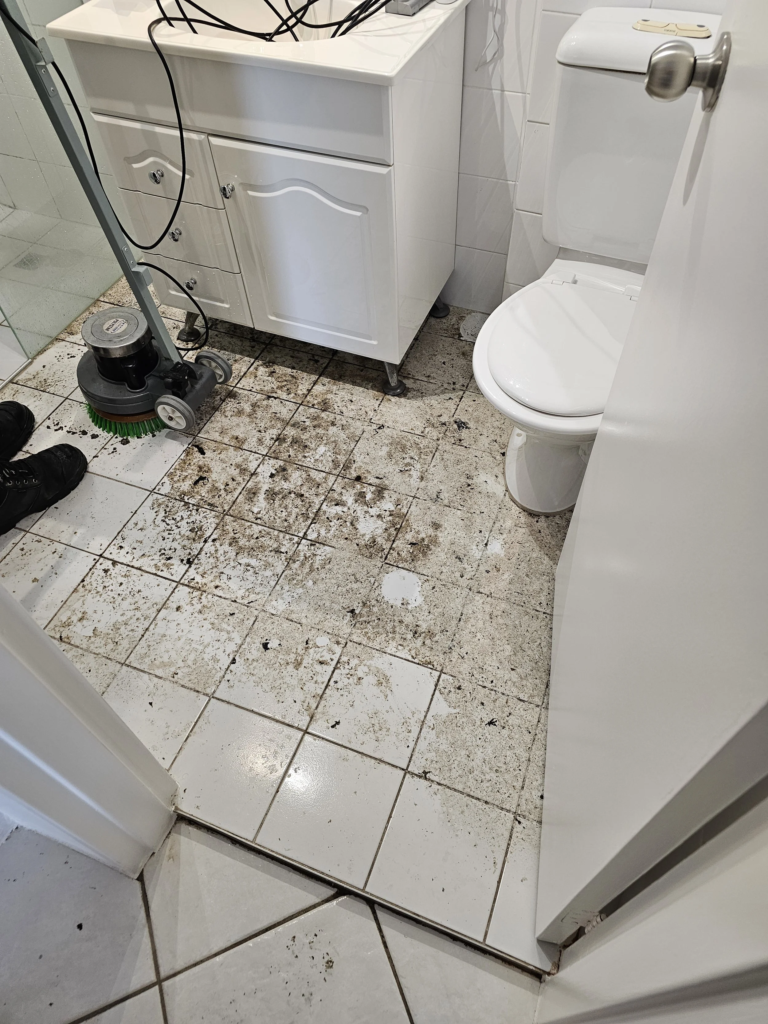 Bathroom floor covered with dirt and debris, surrounding the base of a white cabinet and toilet. There is a janitorial floor cleaning machine in the lower left corner, and black cords on top of the cabinet.