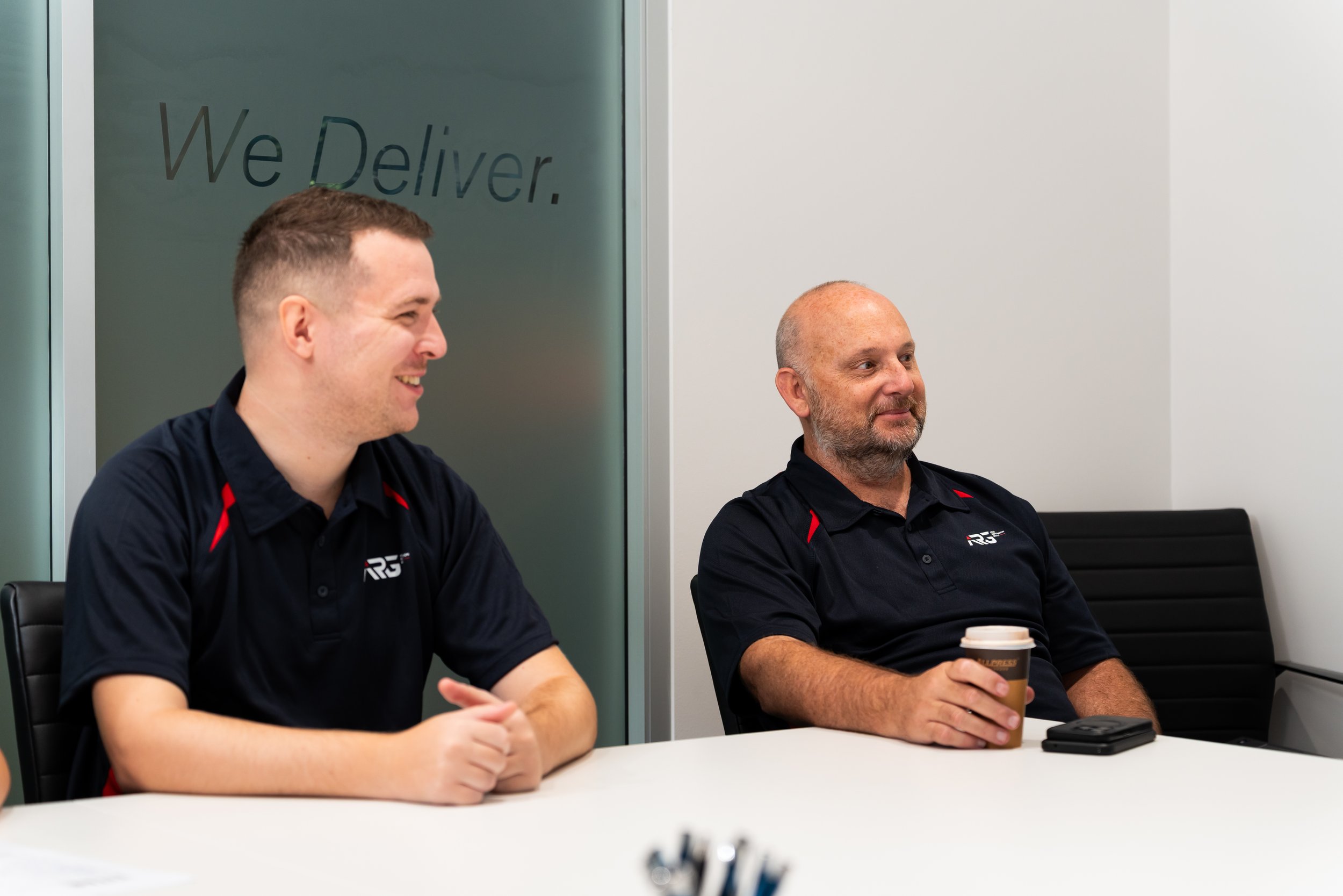 Two men sitting at a conference table in a modern office, wearing black shirts with a logo, one smiling and the other holding a coffee cup.