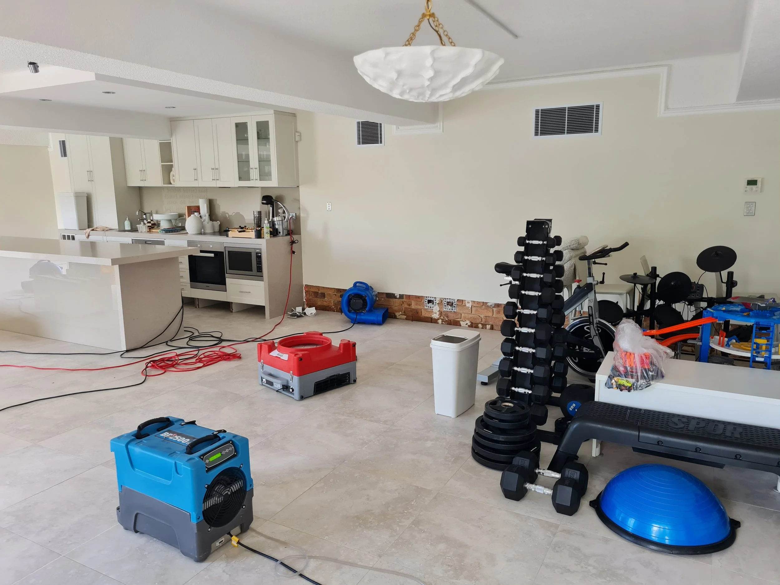 A kitchen room with white cabinets and a variety of home gym equipment including a weight rack, exercise balls, and a blue ventilation fan on the floor, with some tools and supplies scattered around.