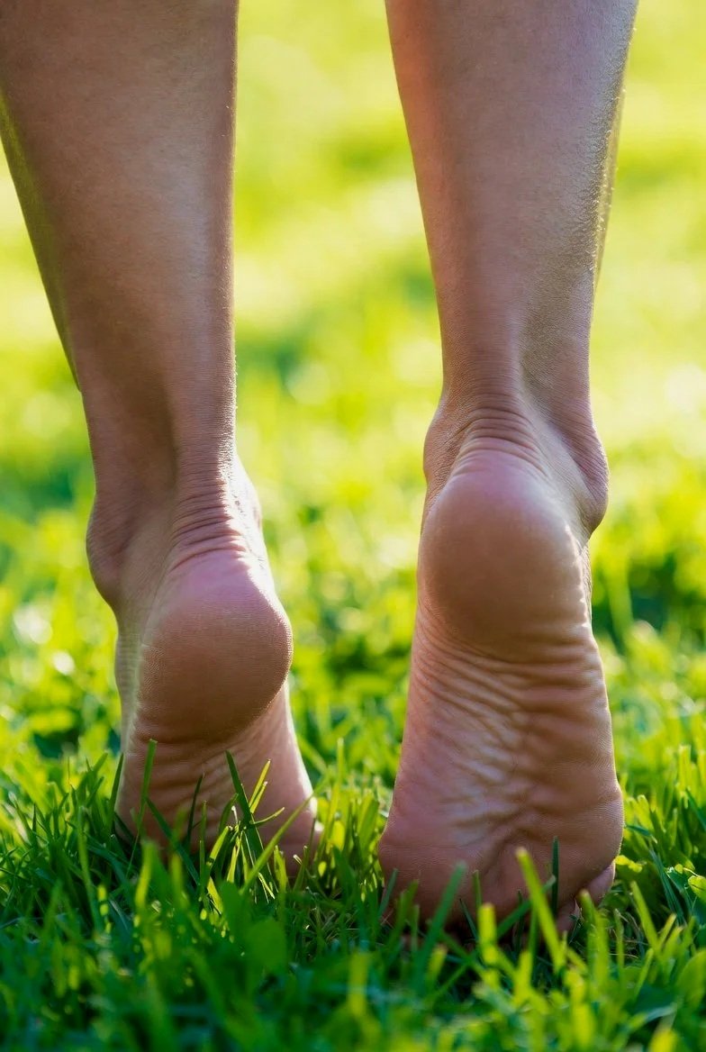 Close-up of bare feet walking on green grass outdoors, with blurred background of sunlight and foliage.