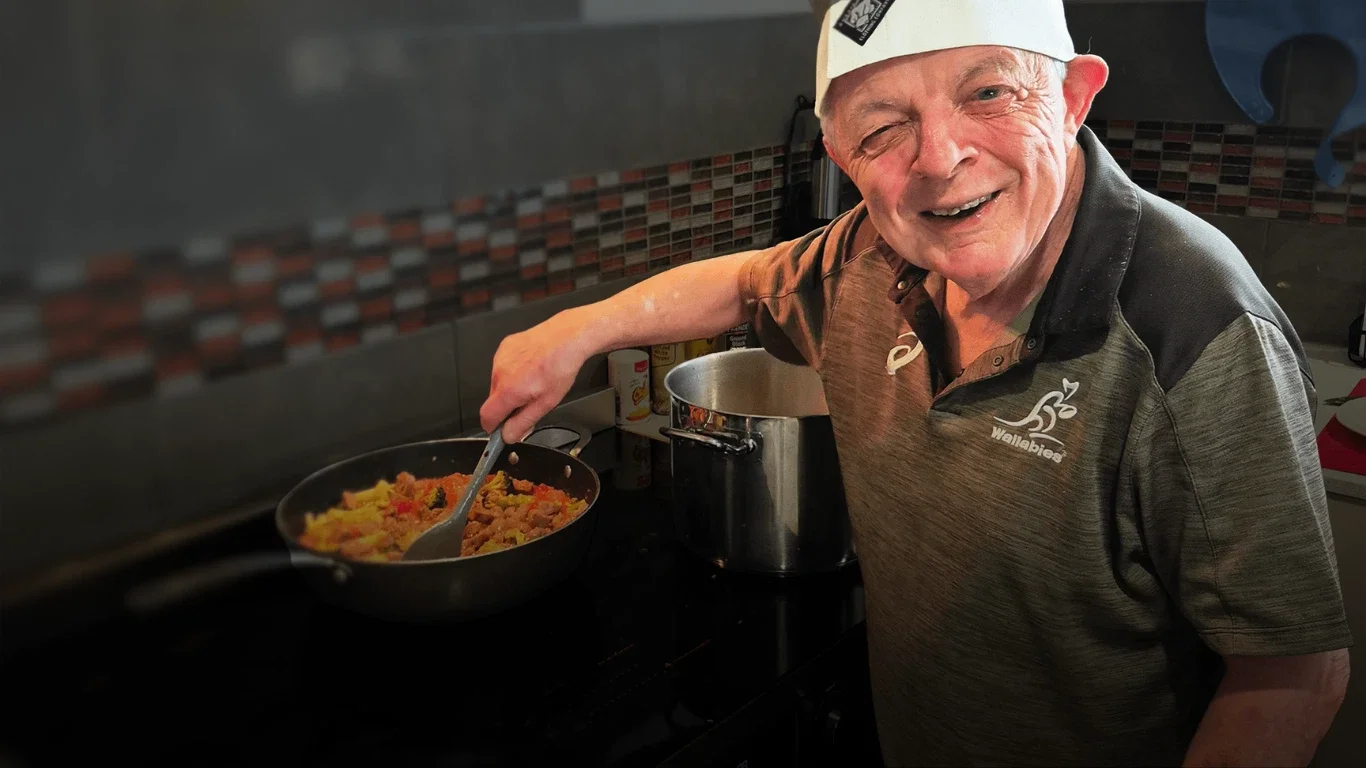 Man cooking food on stove, smiling, wearing a wallabies shirt and a white cap.