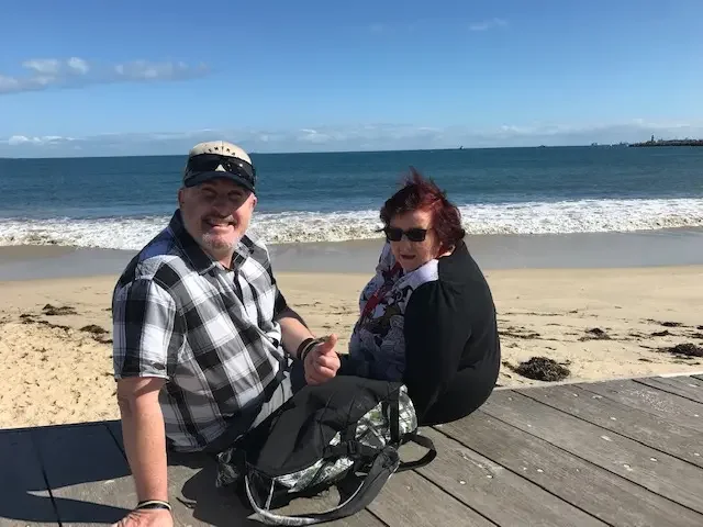 A man and a woman sitting on a wooden bench near the beach with ocean waves in the background.