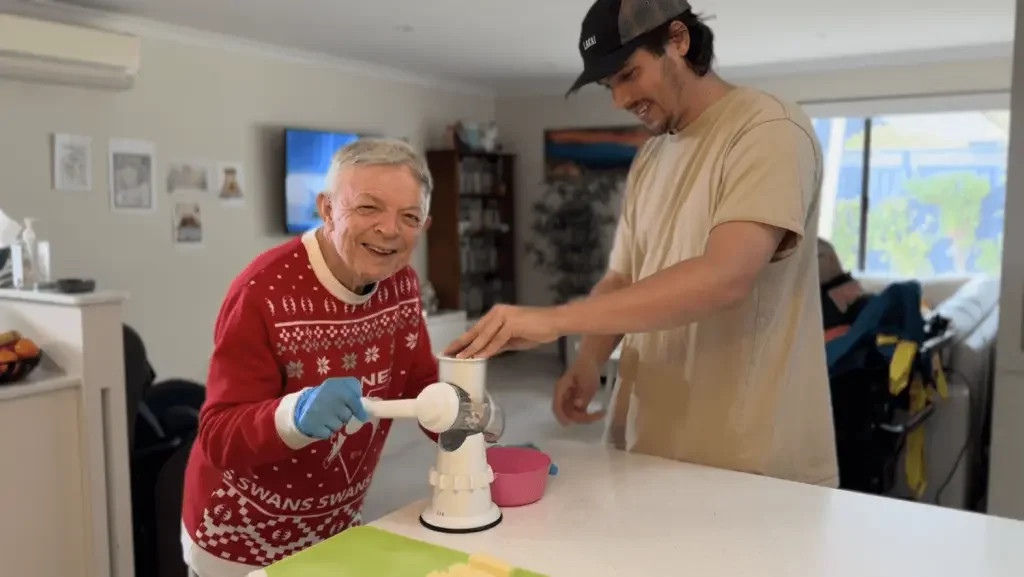 Older man in a red Christmas sweater and blue gloves smiling as he operates a kitchen appliance, with a young man next to him wearing a beige t-shirt and black cap, in a well-lit room with a window and TV in the background.