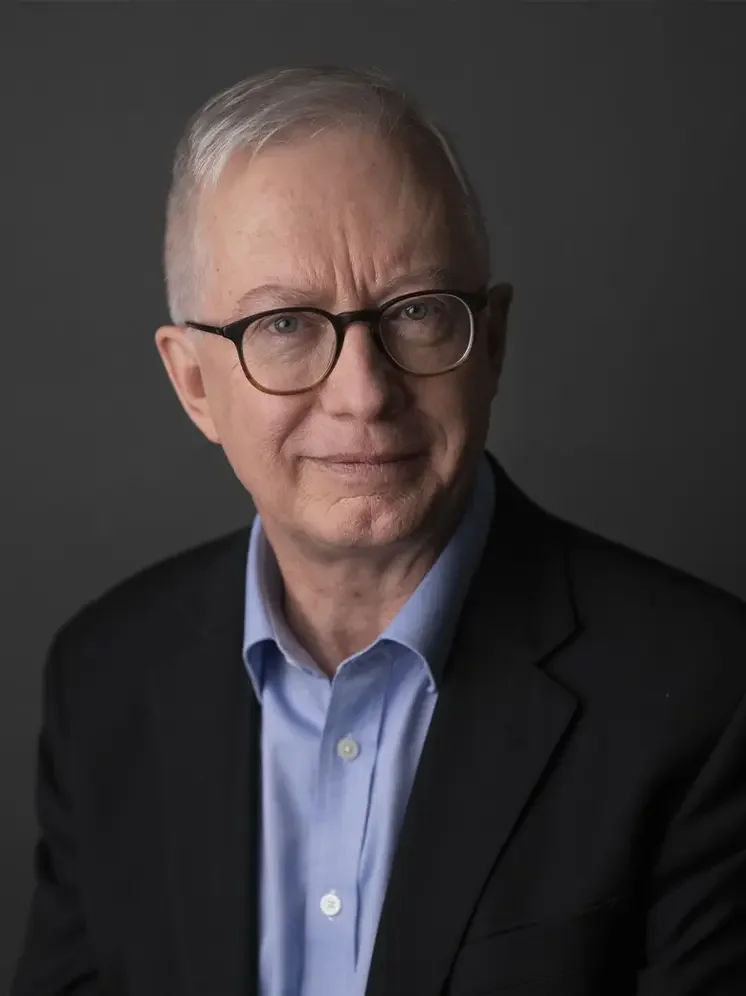 Portrait of an older man wearing glasses, a blue shirt, and a dark blazer against a dark background.