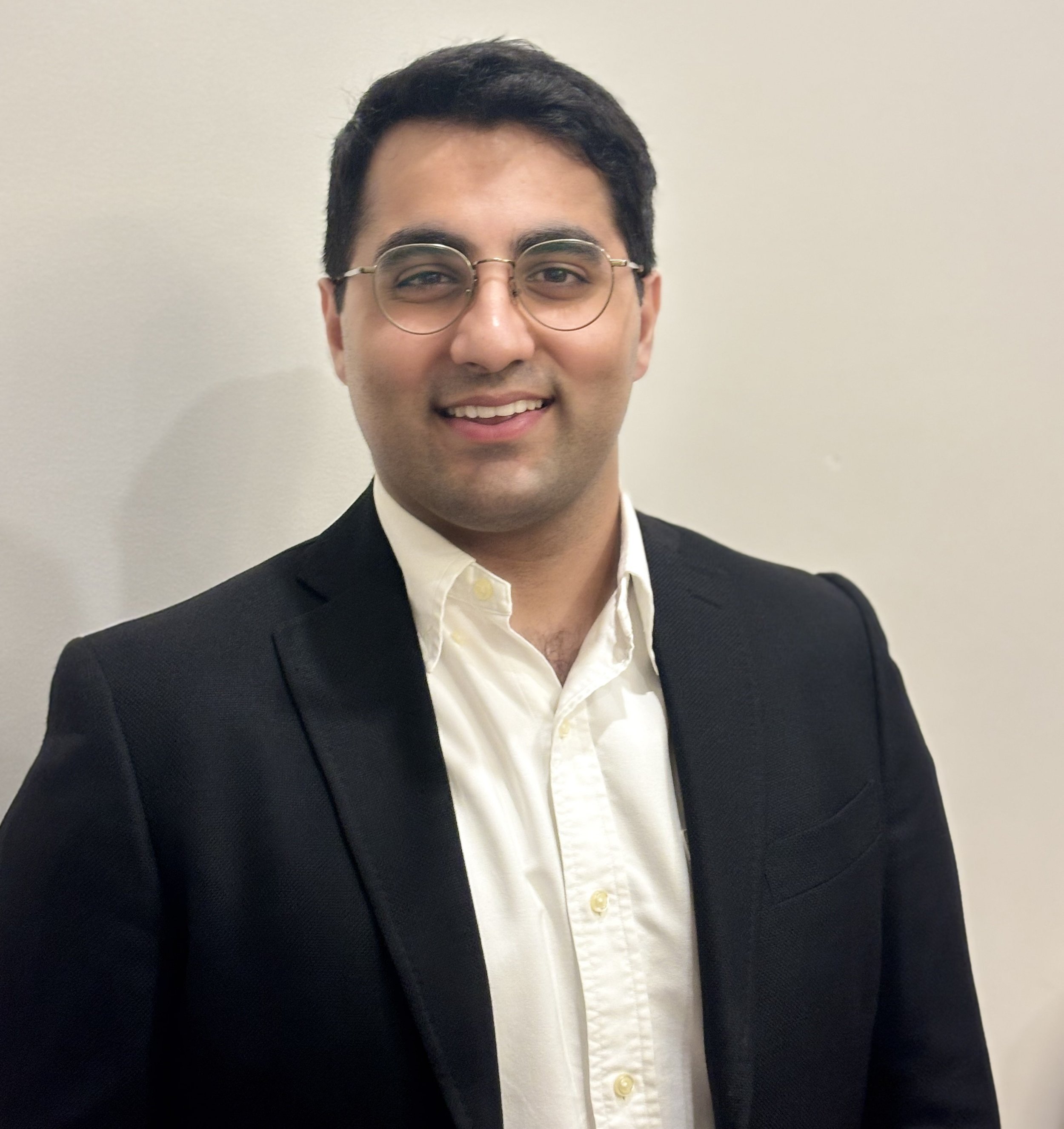 A young man with dark hair, wearing glasses and a black blazer, standing against a plain beige wall, smiling at the camera.