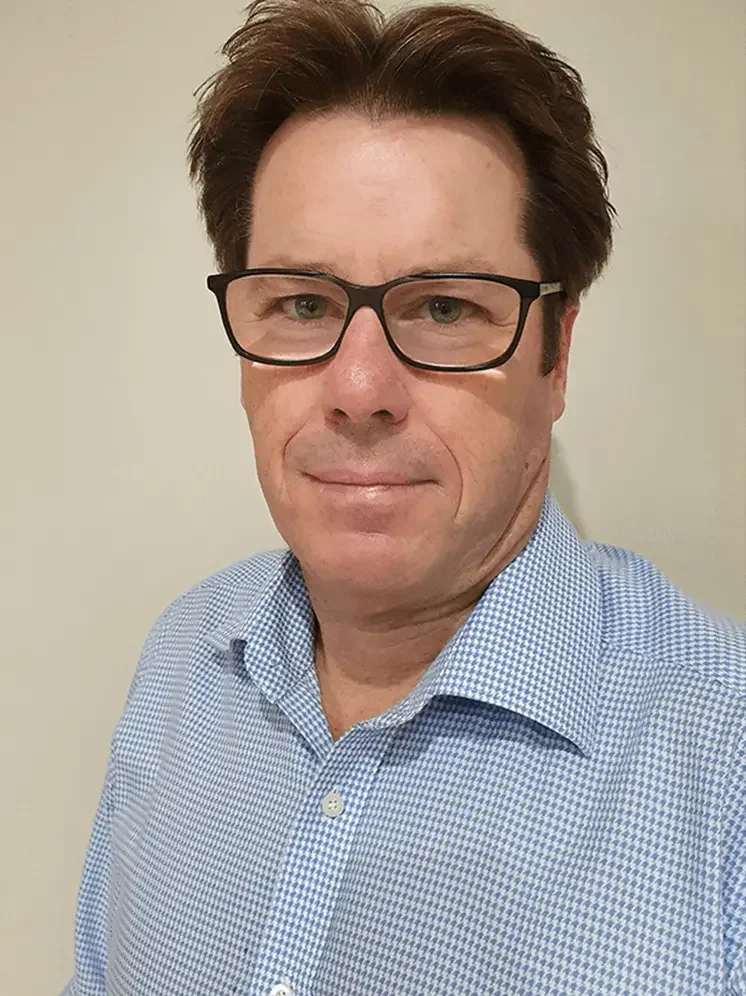 A middle-aged man with short brown hair, wearing glasses and a blue checkered shirt, facing the camera with a neutral background.