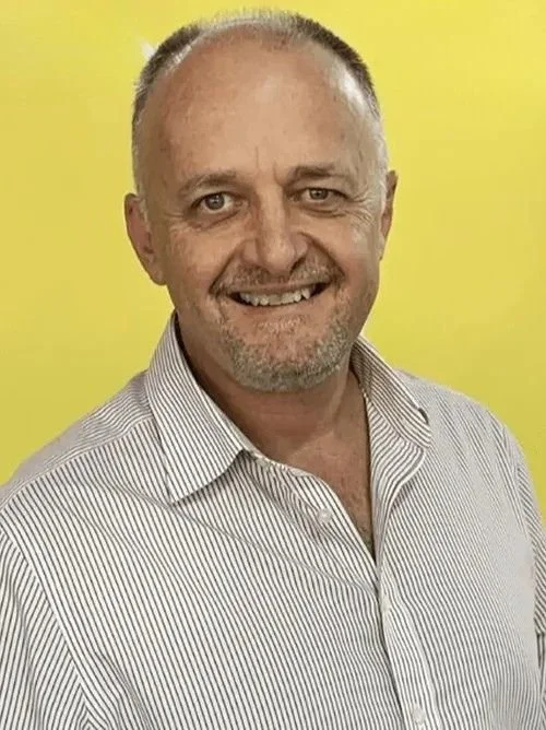 A middle-aged man smiling, wearing a striped dress shirt, with a yellow background.