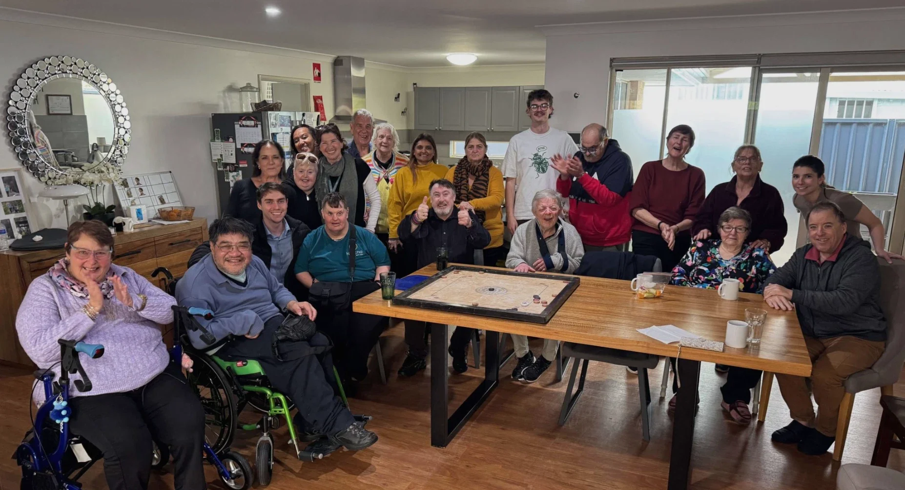 Group of people gathered around a table in a bright room, celebrating a social event or game night, with some smiling and clapping.