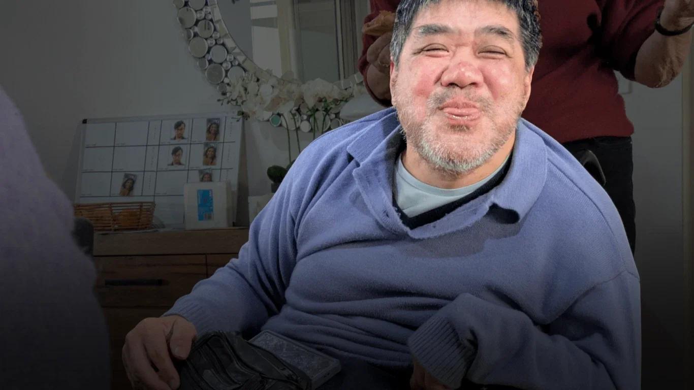 A man with gray hair and a beard making a funny face, sticking out his tongue, while sitting in a room with a mirror, floral decorations, and photos on display in the background.