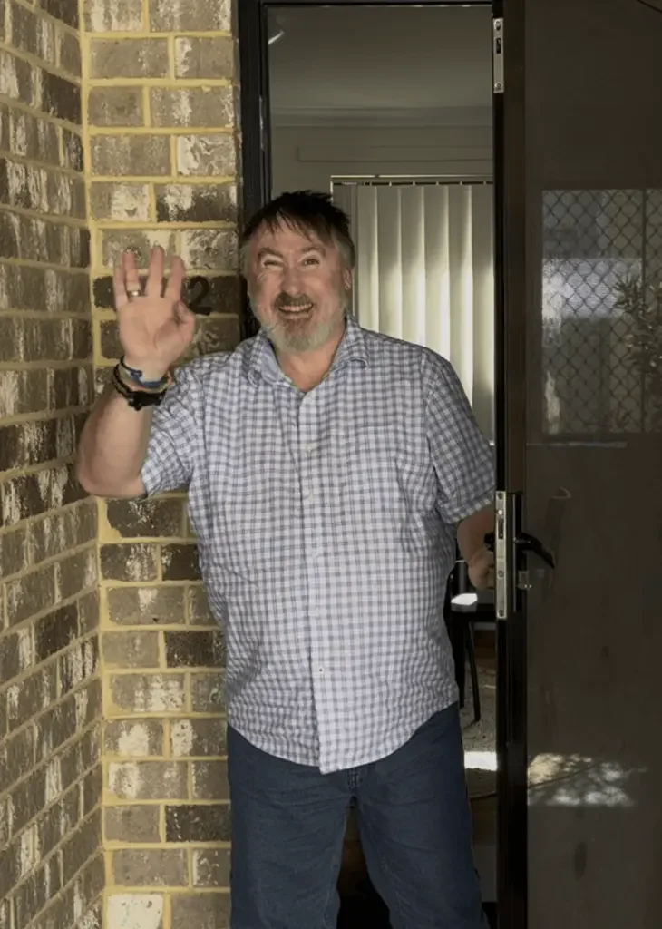A man with gray hair and beard waving and smiling, standing at an open door outside a house. The man is wearing a checkered shirt and dark pants, and the background shows a window with vertical blinds and a screened door.