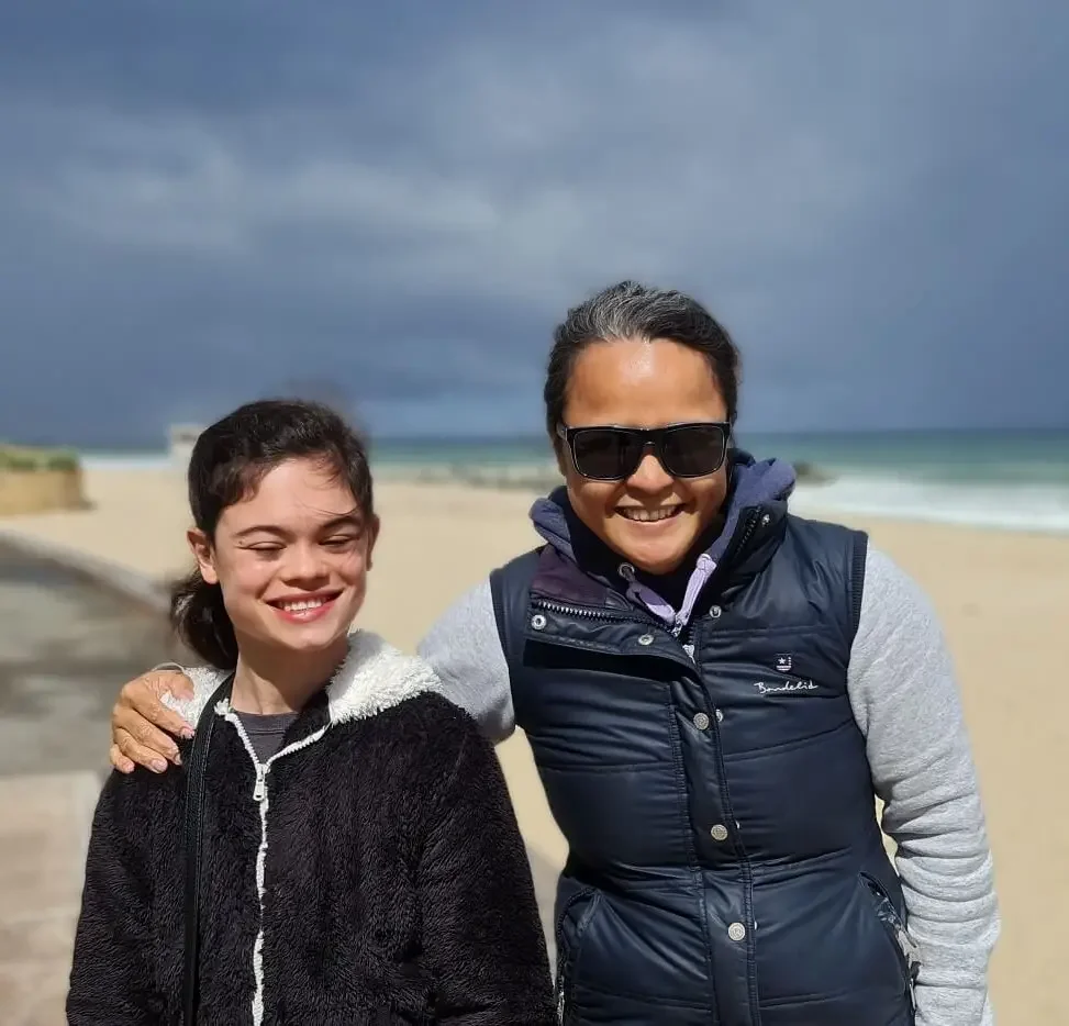 A smiling woman and girl at the beach on a cloudy day, with the ocean in the background.