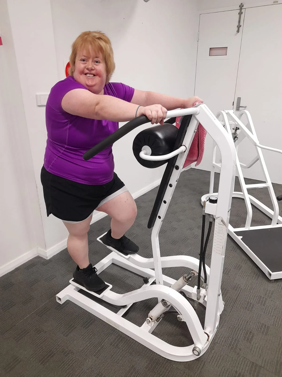 A woman with short red hair smiling while exercising on a stepper machine in a gym or therapy room.