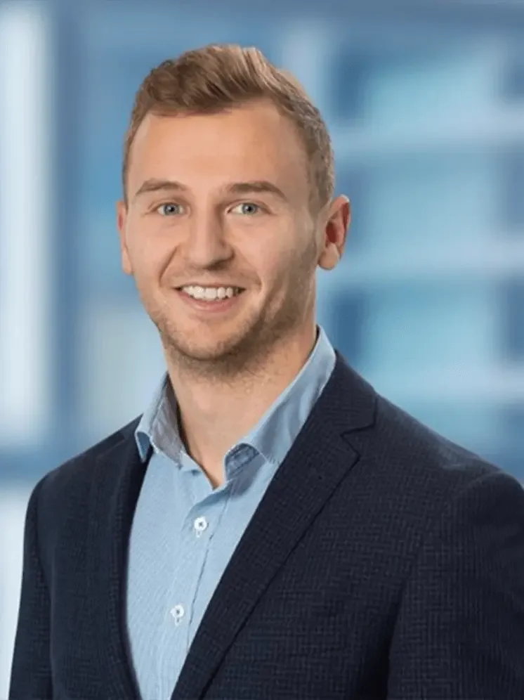 A young man with light brown hair, wearing a navy blazer and light blue collared shirt, smiling in front of a blurred blue-toned office background.
