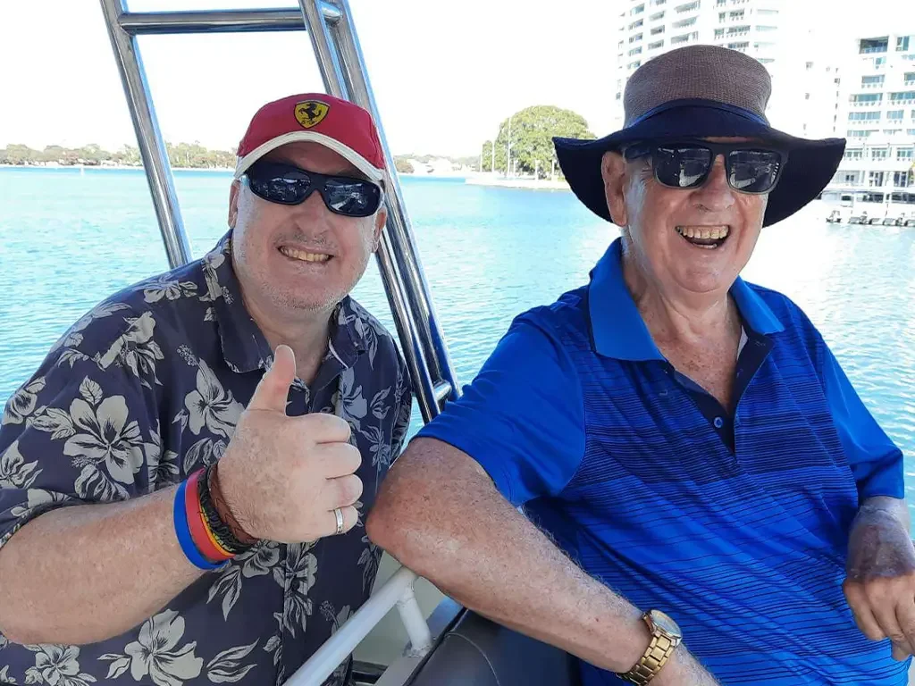 Two smiling men wearing sunglasses, one giving a thumbs-up, on a boat with water and buildings in the background.