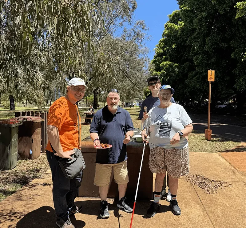 Four men standing outdoors near a picnic table, with trees and a clear blue sky in the background. One man is holding a plate of food, another has a walking cane, and they are all smiling.