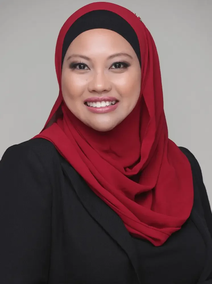 A woman with a hijab, wearing a red headscarf and black blazer, smiling at the camera against a gray background.