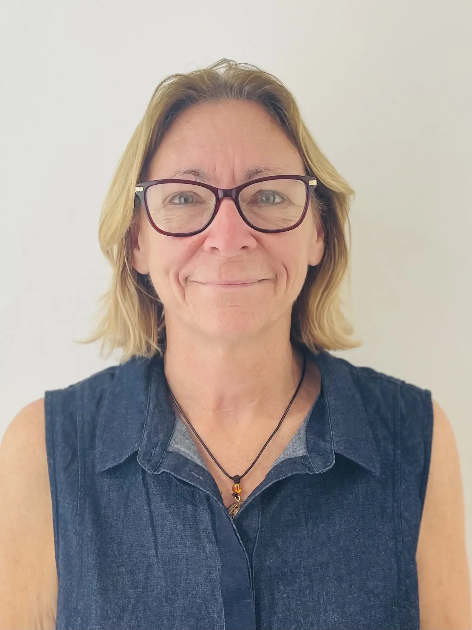 Close-up of a woman with shoulder-length blonde hair wearing glasses, a black sleeveless shirt, a grey undershirt, and a necklace with a bead pendant, smiling against a plain white wall.