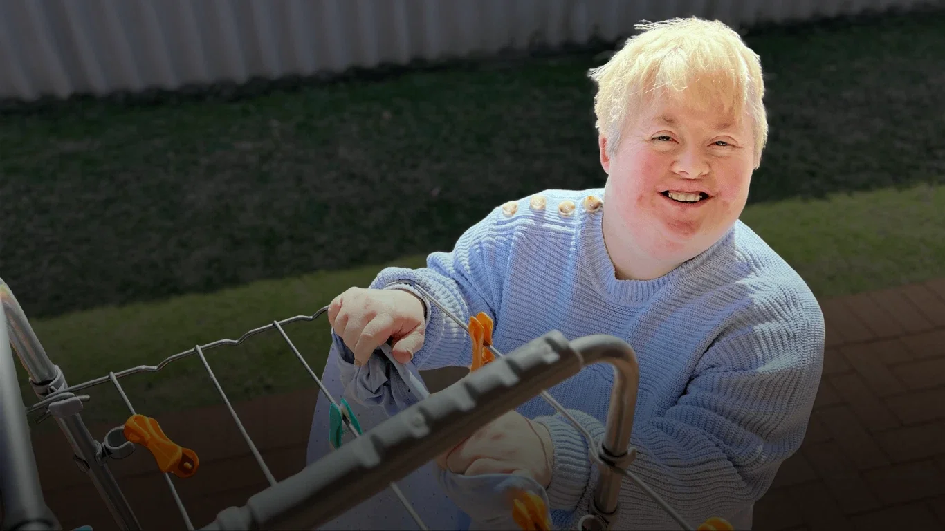 A young person with blonde hair smiling and hanging laundry with clothespins on a clothes drying rack outside.