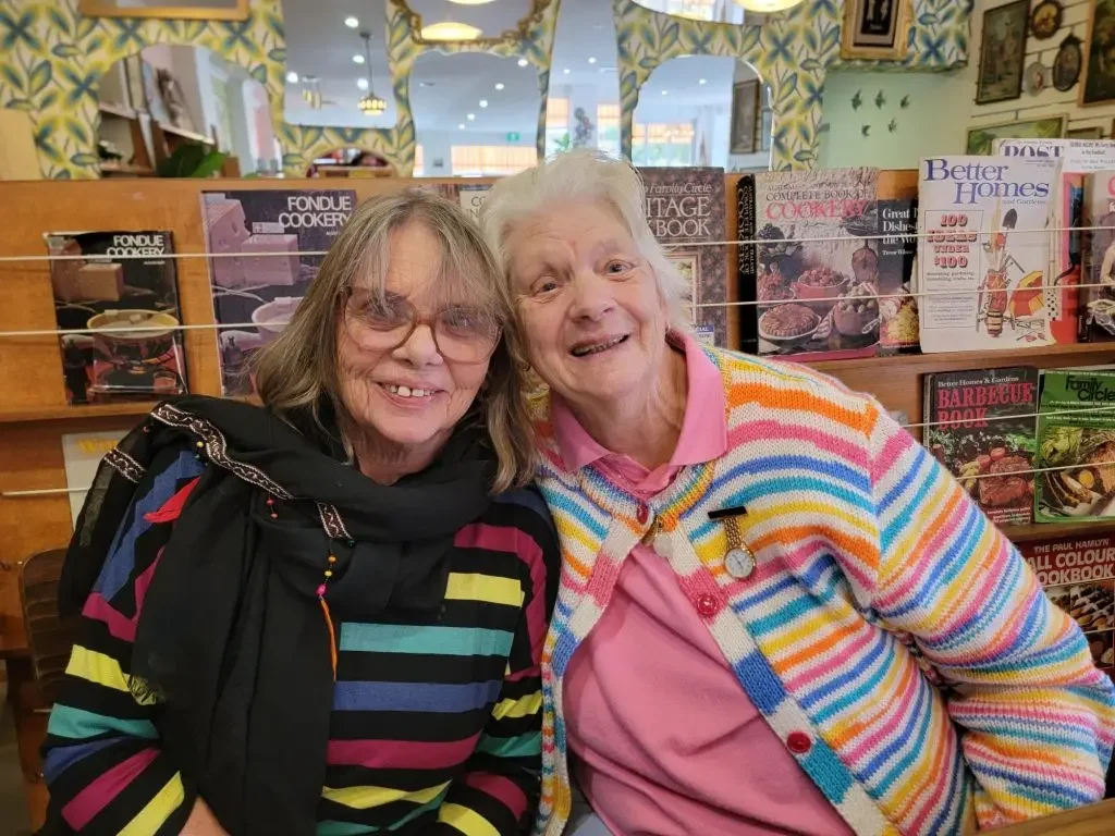 Two women smiling and posing together inside a bookstore with colorful magazine racks and patterned wallpaper in the background.