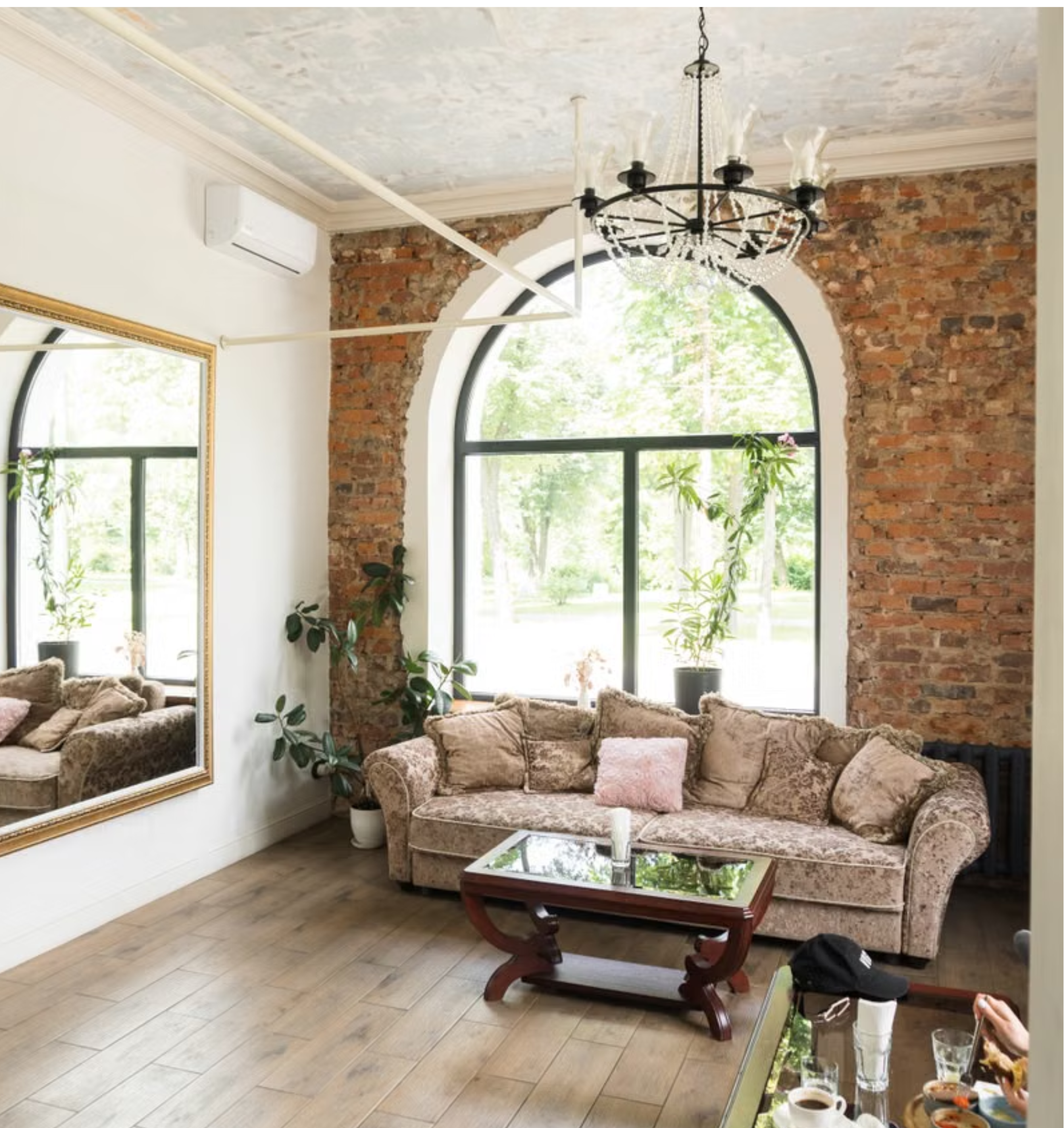 Living room with large arched window, brick wall, vintage floral sofa, glass-top coffee table, plants, and chandelier.