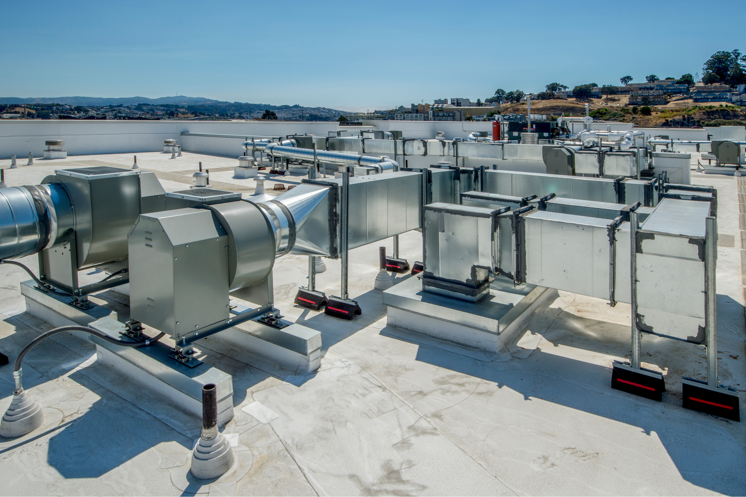 Rooftop HVAC system with metal ductwork, vents, and equipment against a bright blue sky, with distant hills and residential buildings on the horizon.