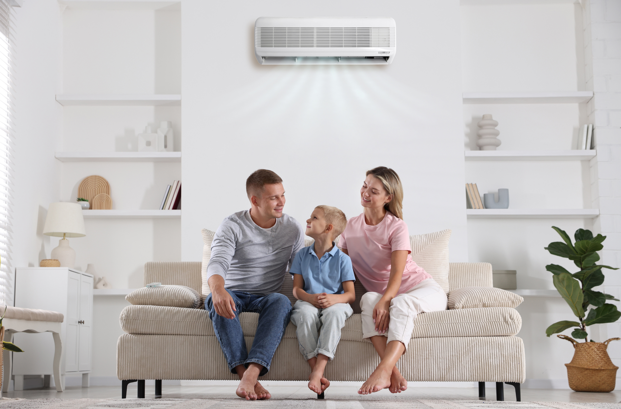 A family of three sitting on a beige striped sofa in a bright living room, with a white wall-mounted air conditioner above them. The family includes a man, a woman, and a young boy sitting between them, holding hands and smiling at each other. The room has white shelves with decorative items, a lamp, and a potted plant.