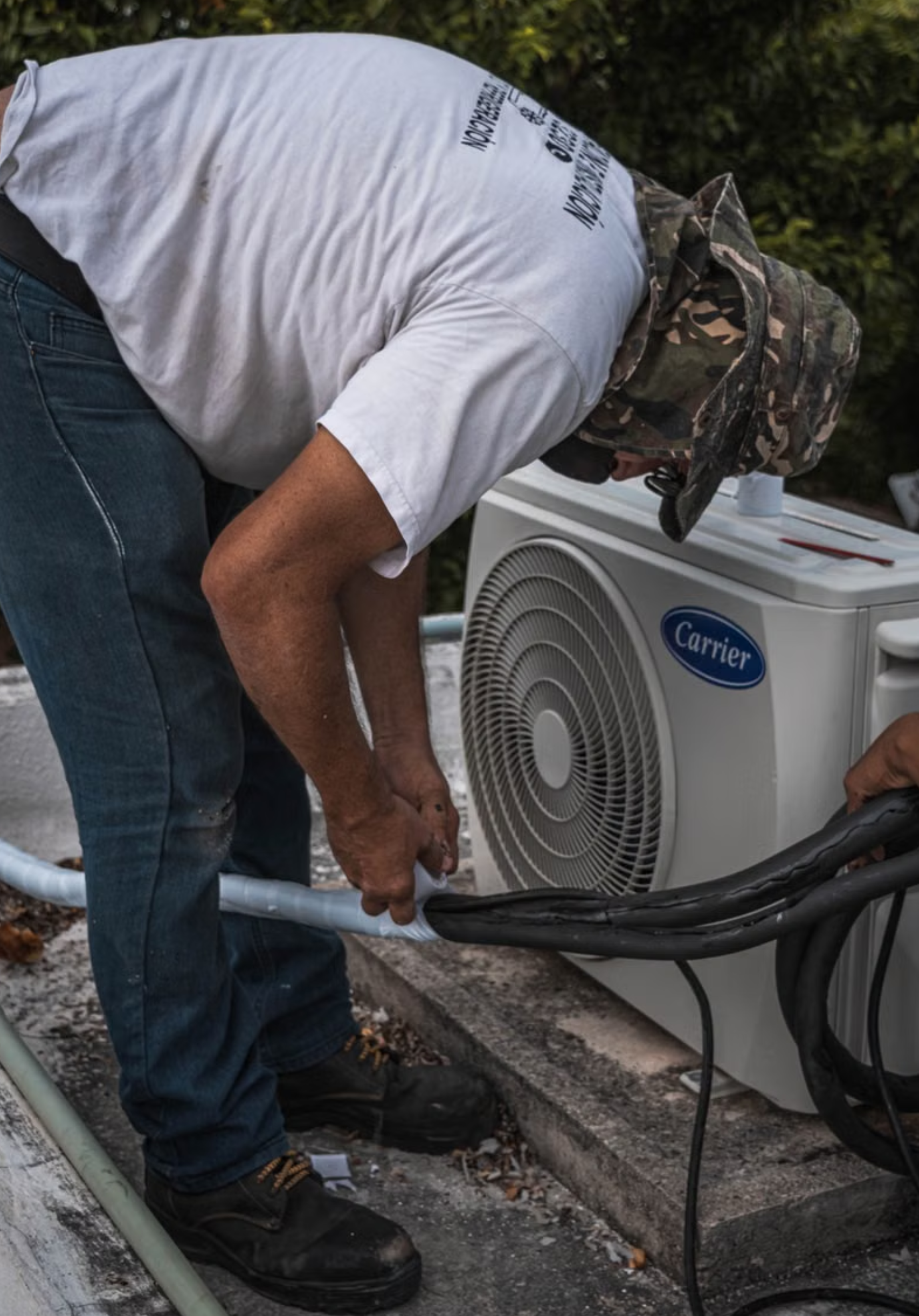 A person wearing a white t-shirt and camouflage hat working on outdoor air conditioning unit.