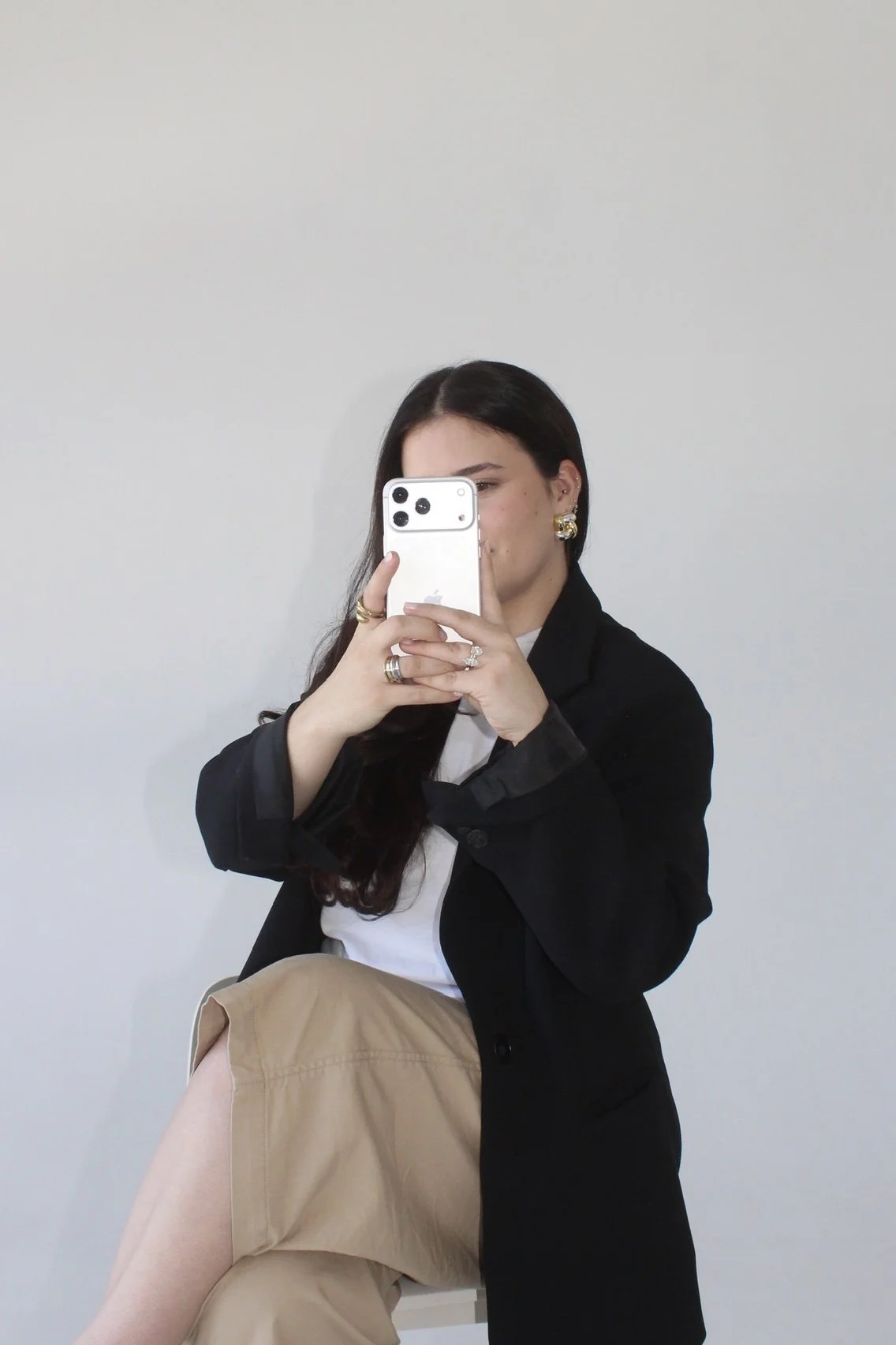 A woman taking a mirror selfie with her smartphone, wearing a black blazer, white shirt, beige pants, and gold jewelry, against a plain white wall.