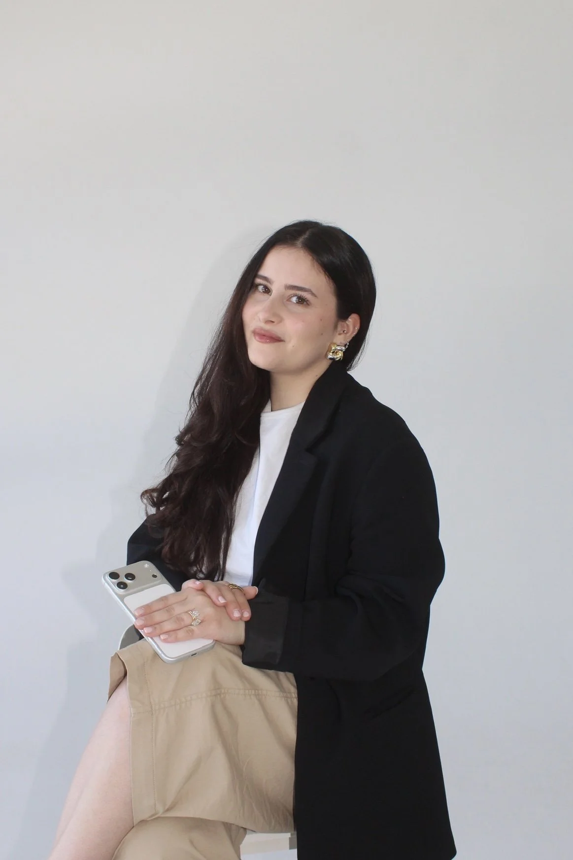 A woman with long dark hair, wearing a black blazer and white shirt, sitting against a plain light-colored wall, holding a smartphone, with her leg crossed over her knee.