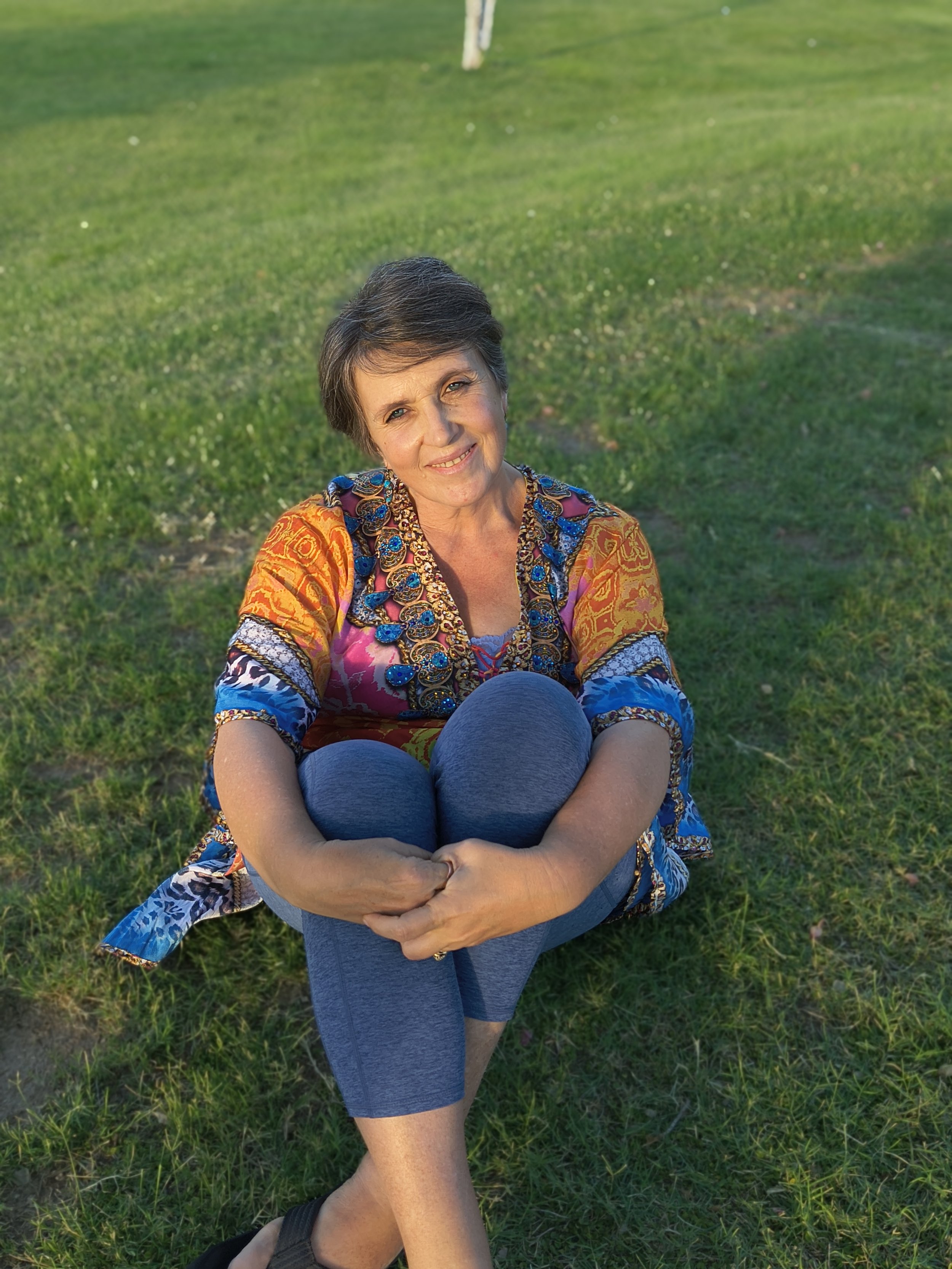 A woman smiling while sitting on the grass in a park, wearing a colorful patterned blouse and gray leggings, with her right leg crossed over her left knee.