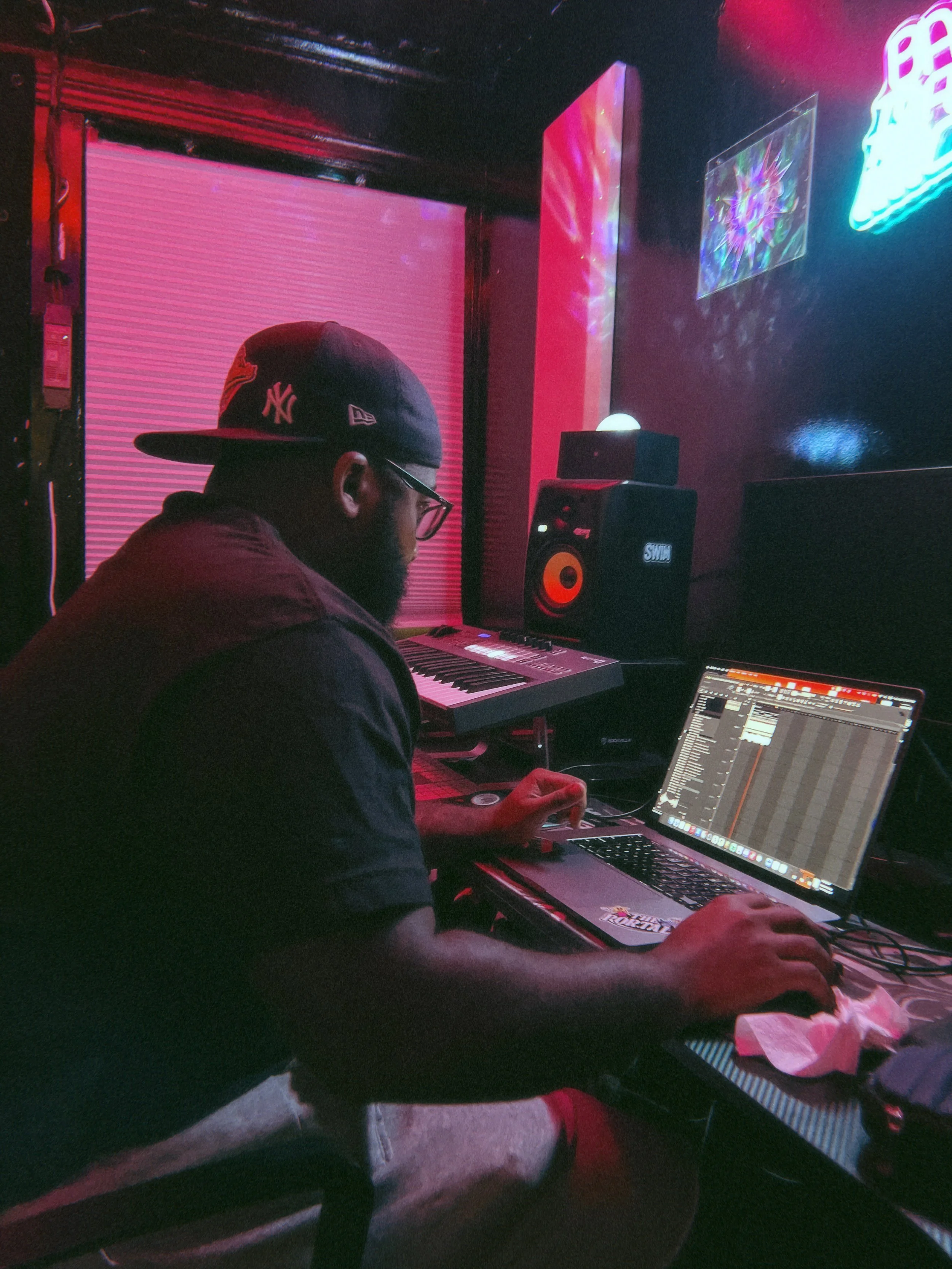 A person wearing a black New York Yankees cap and glasses is working on a music production setup with a laptop, MIDI keyboard, and studio speakers in a dimly lit room. Neon and LED lights cast colorful reflections on the wall.