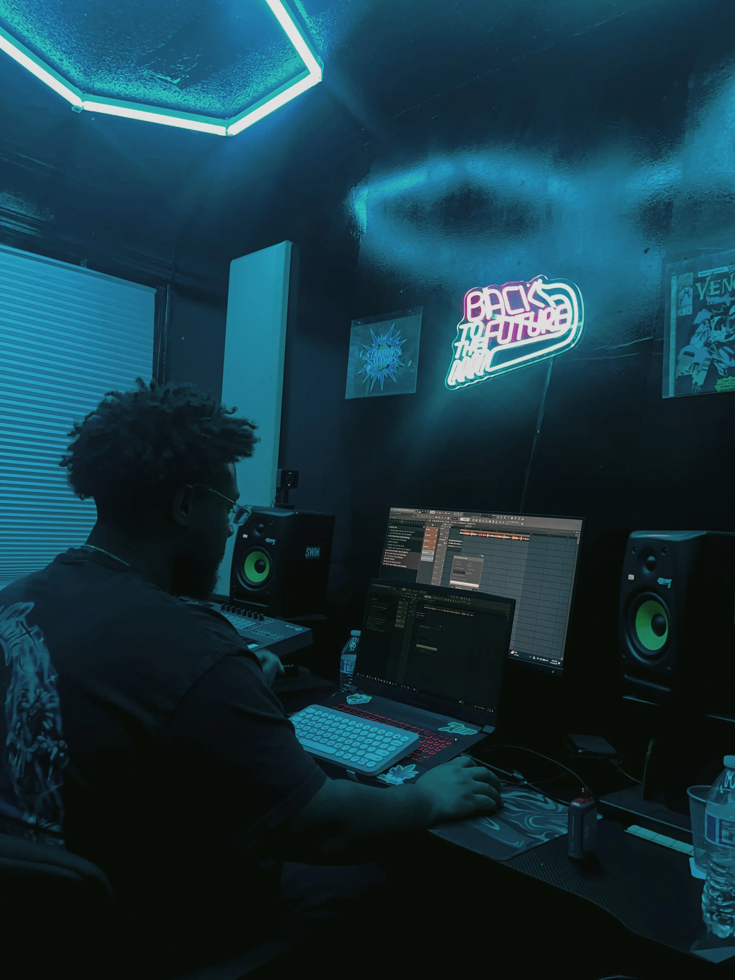A person working on music production at a computer desk in a dimly lit room with dark walls, neon signs, and posters on the wall.