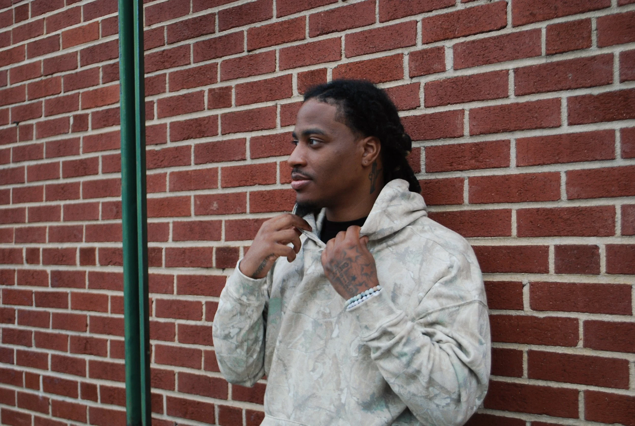 A young man with dark hair in dreadlocks and tattoos on his hands and neck, wearing a light patterned hoodie, standing against a red brick wall. He is looking to his right and holding the hoodie's drawstrings with both hands.