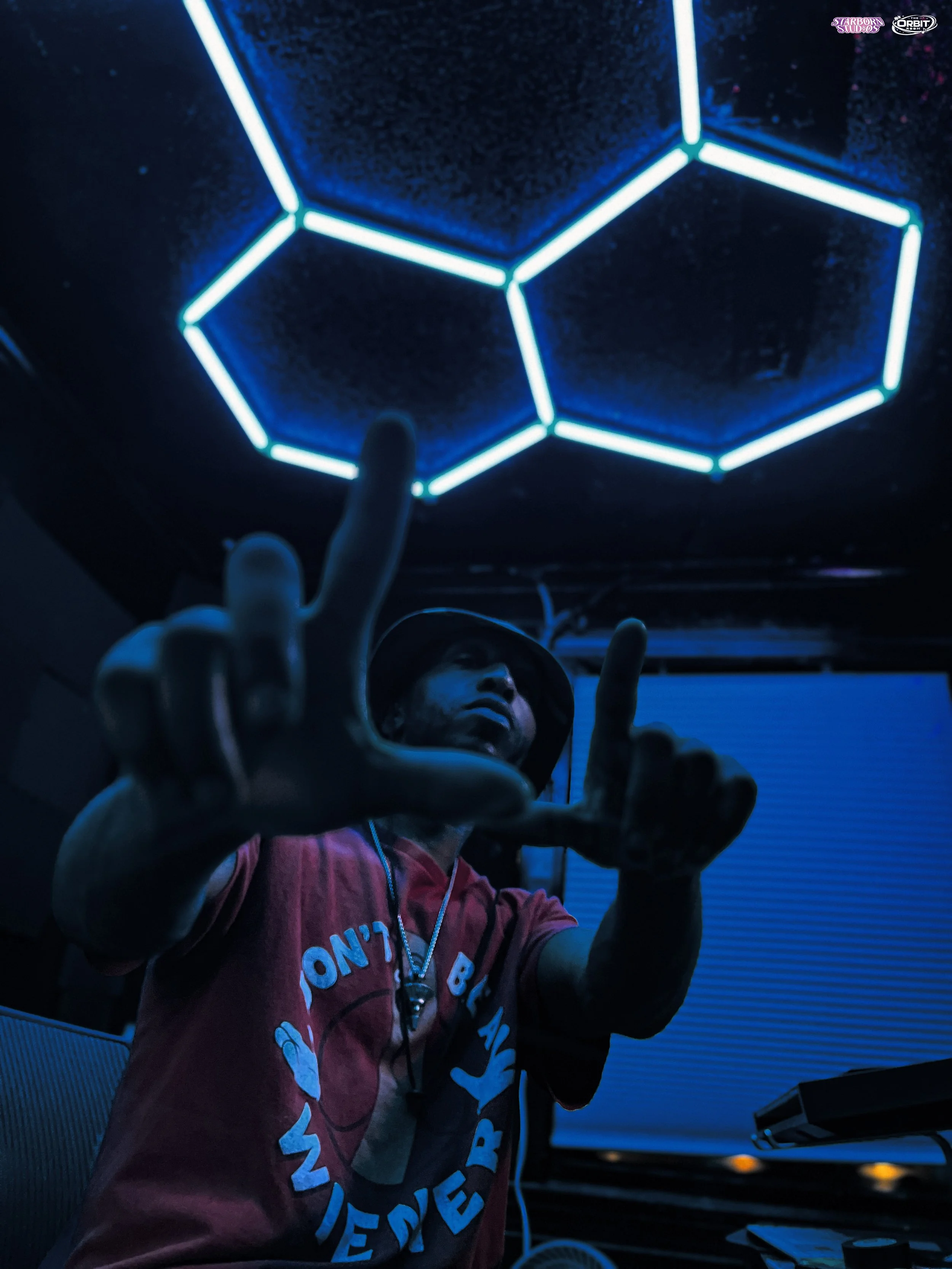 A person wearing a red shirt and a wide-brimmed hat pointing at neon-lit hexagons on a dark ceiling, with blue lighting and a computer monitor in the background.