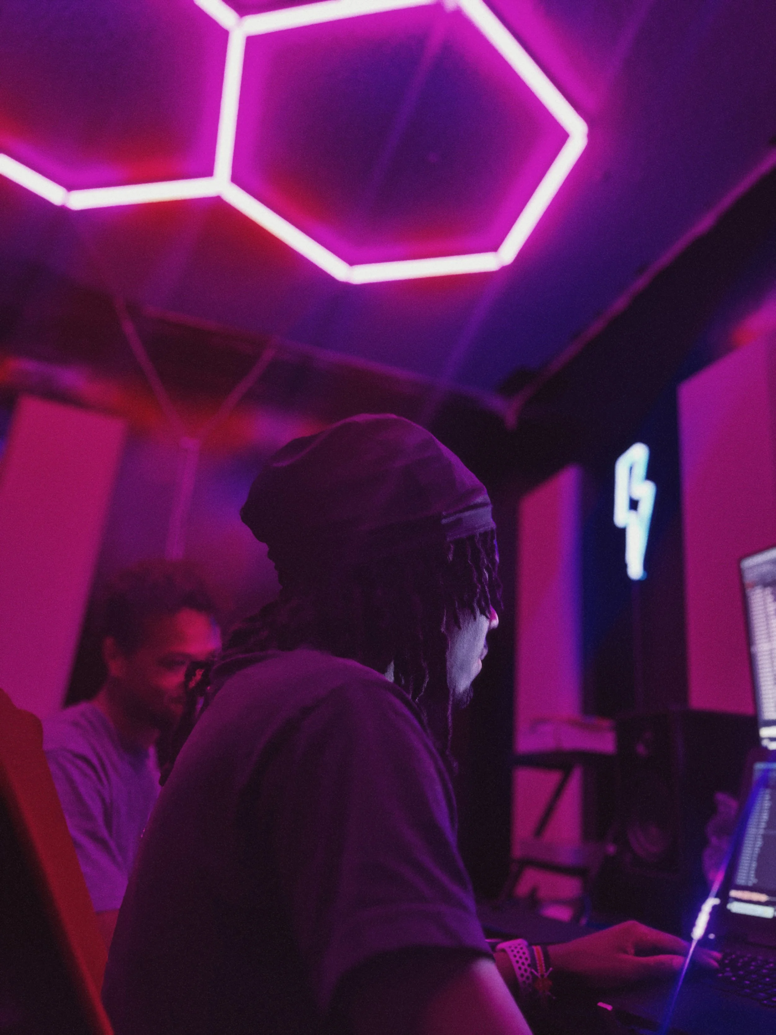 Two young men seated in a dimly lit room with purple and pink neon lighting, focused on their computer screens.