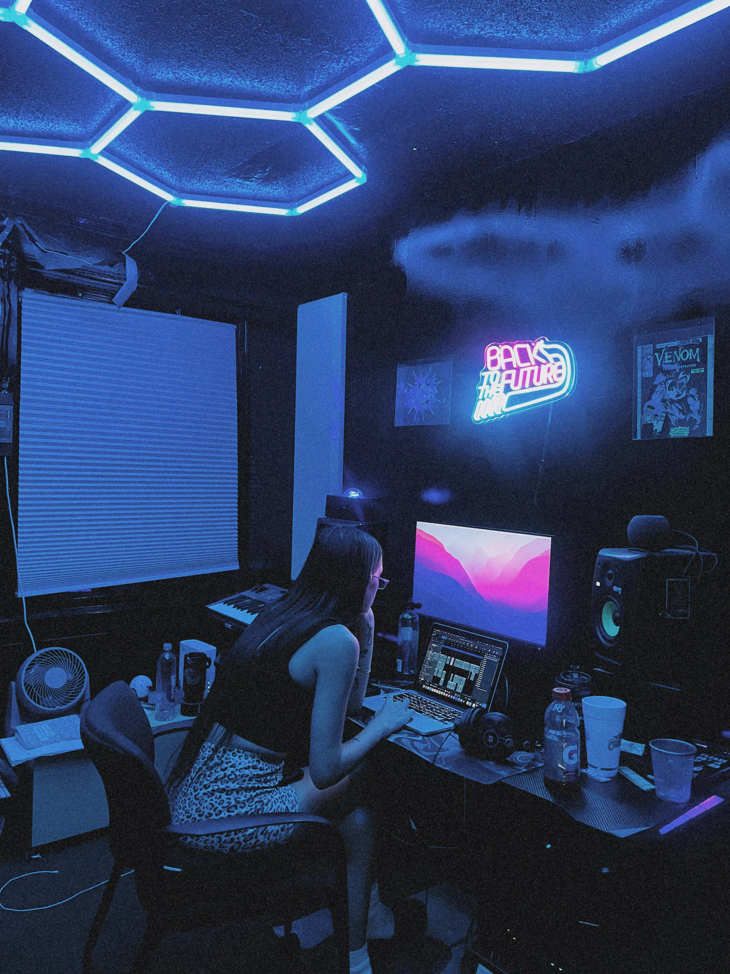 A woman in a leopard print dress and glasses sitting in front of a laptop and music production equipment in a dark room illuminated by neon blue lights. There are water bottles and cups on the table, and a neon sign on the wall that reads 'Back to the Future'.