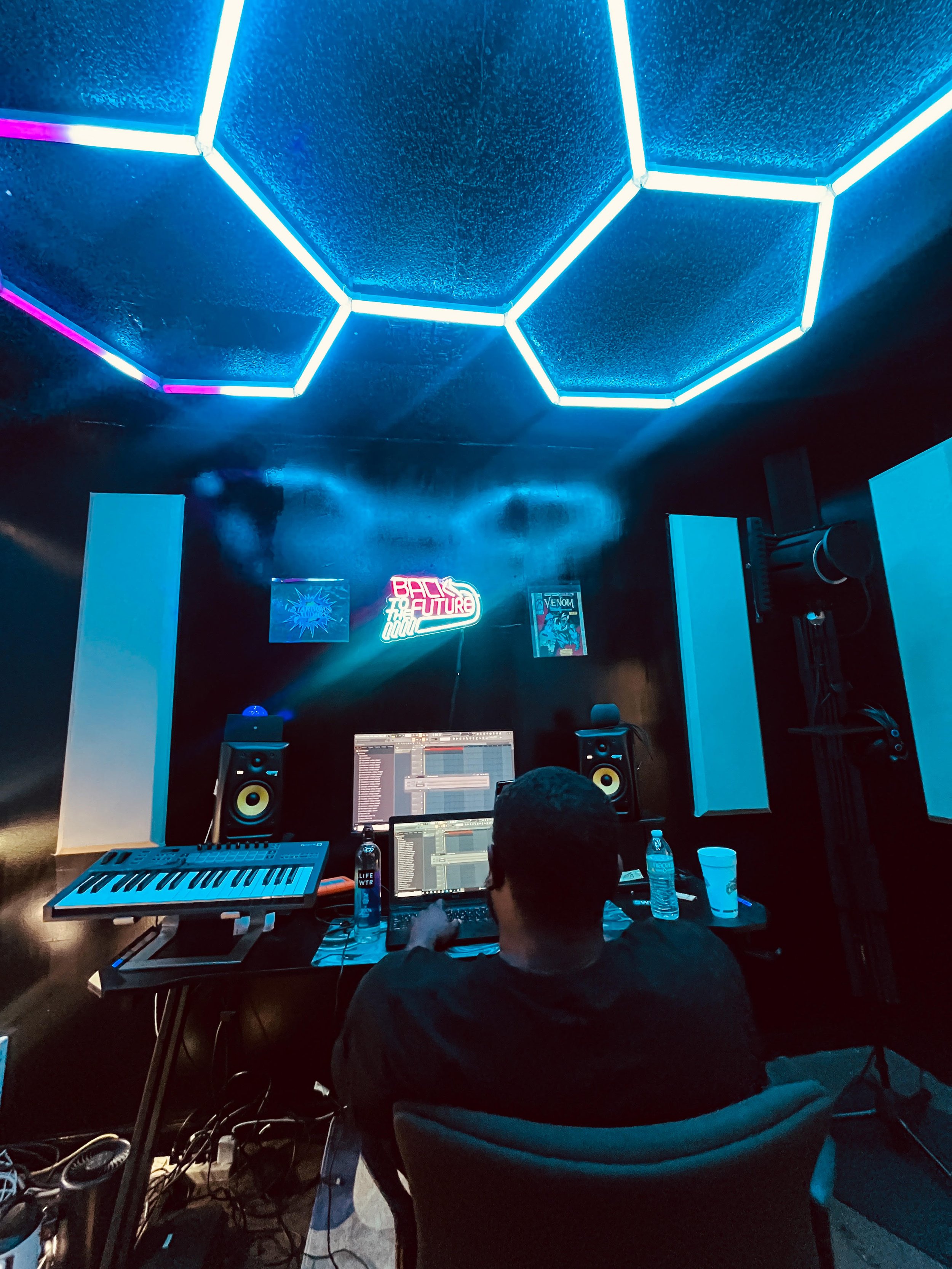 A person sitting in a music studio at a mixing console with computer, keyboard, studio monitors, water bottle, and neon signs on the wall.