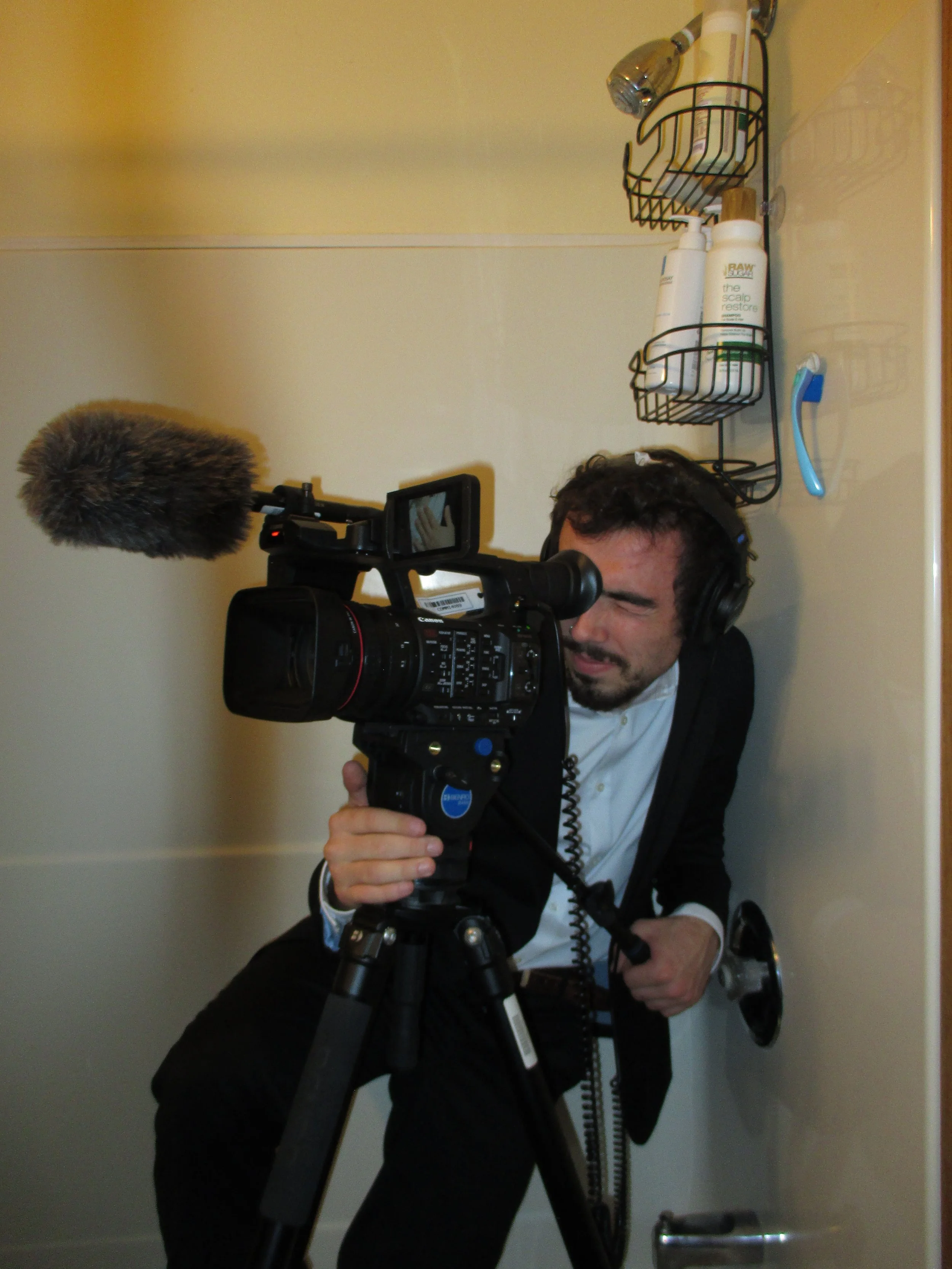 Person with dark hair and facial hair operating a professional video camera with a windscreen microphone, in front of a beige wall with a wall-mounted metal rack holding toiletries, a toothbrush, and a shampoo bottle.