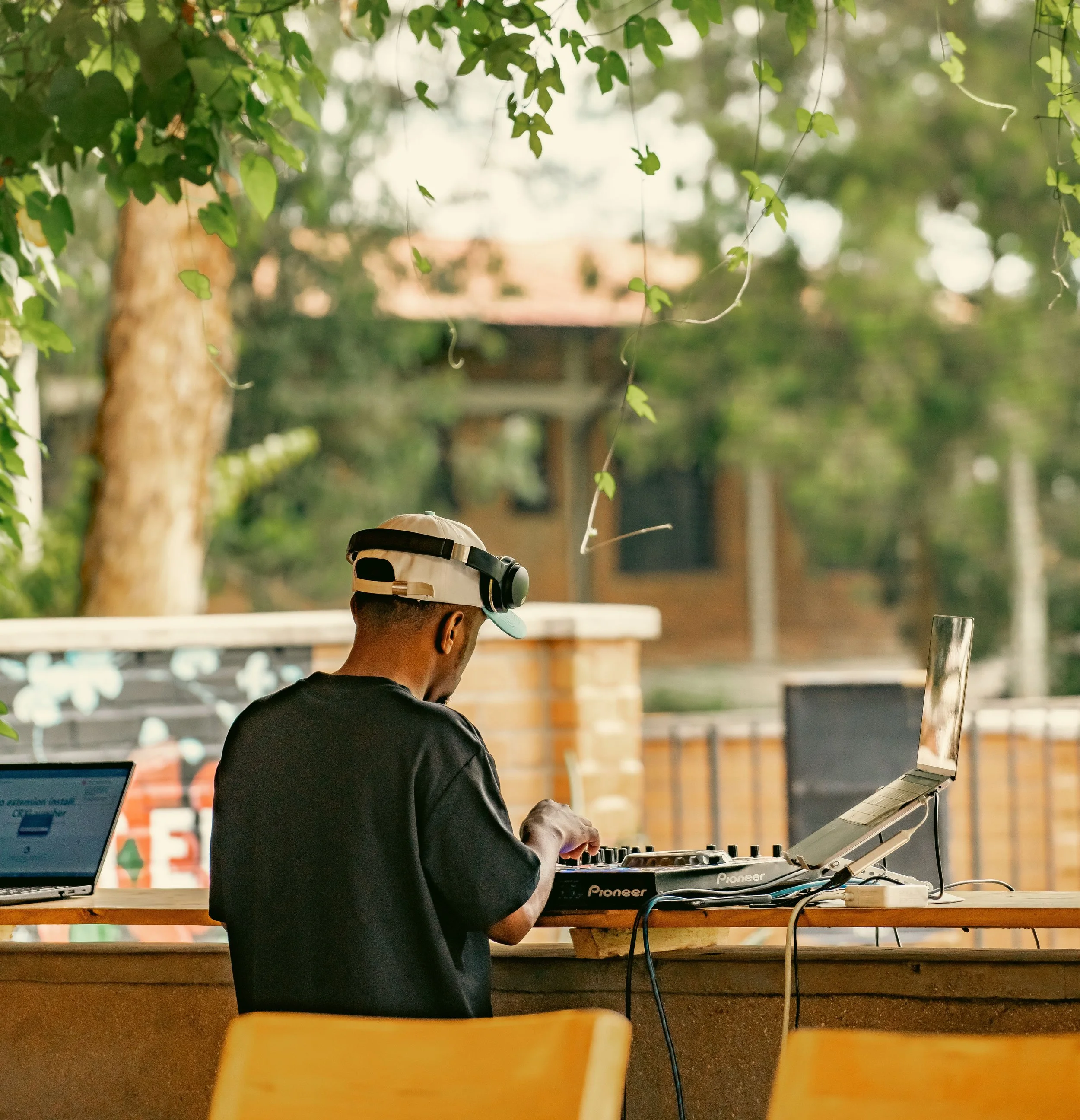 A person wearing a cap operating a DJ mixer in an outdoor setting with green foliage in the background.