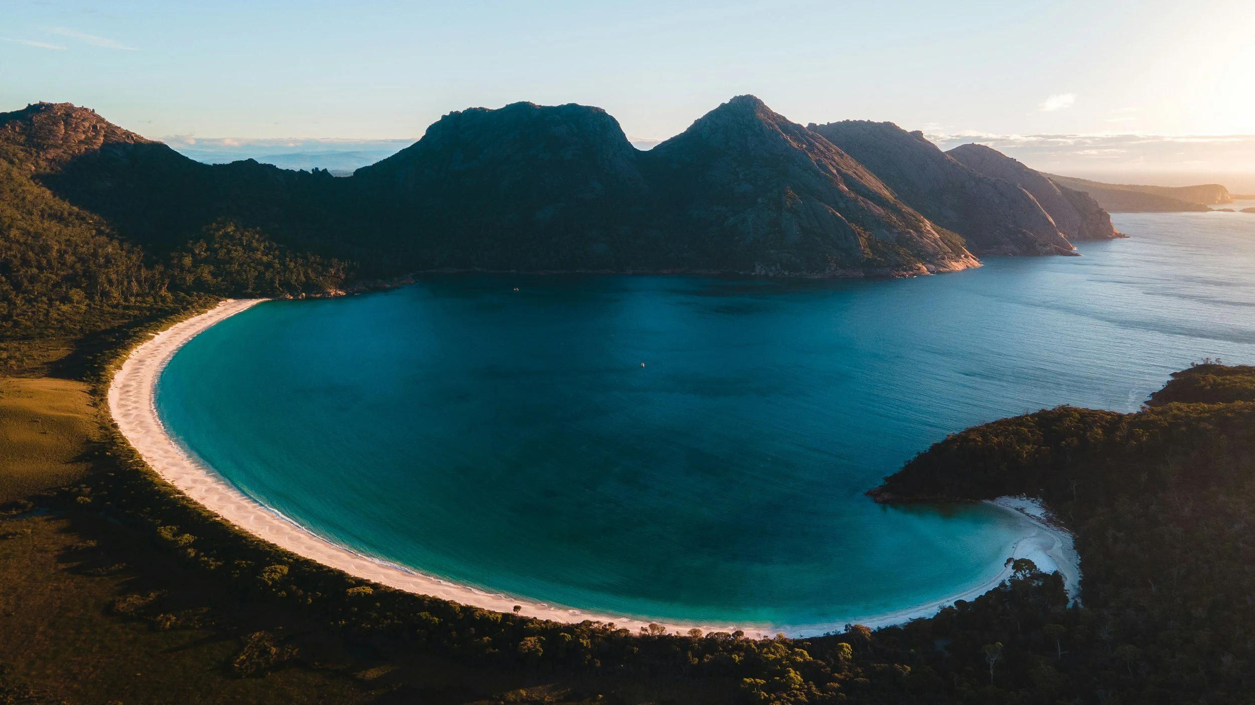 A coastal landscape with a crescent-shaped beach, turquoise water, green forests, and mountainous terrain during sunset.