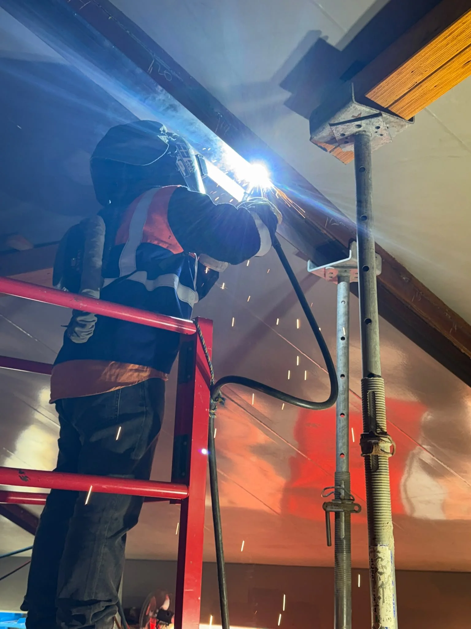Worker in safety gear welding a metal pipe on a scaffold with sparks and bright light in an indoor setting.